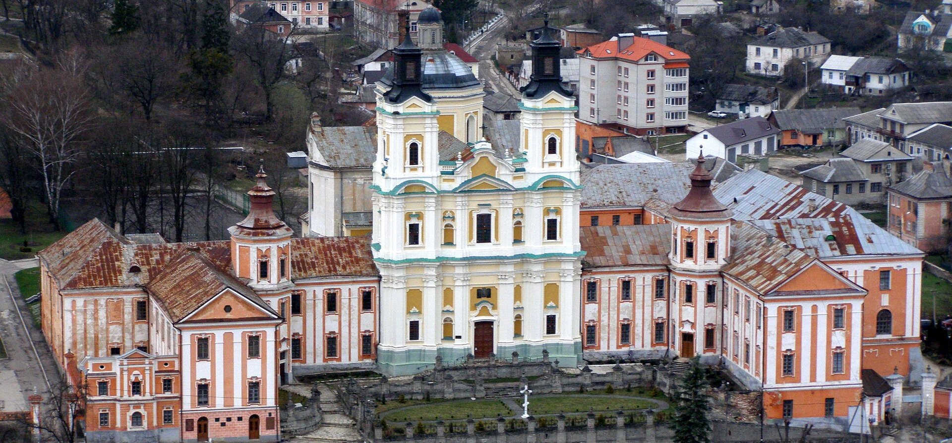 Na zdjęciu znajduje się budynek dawnego liceum w Krzemieńcu. W jego centralnej części znajduje się duży kościół o dwóch wieżach. Ma on jasne ściany i liczne złote zdobienia. Po obu jego stronach rozchodzi się budynek o pomarańczowo żółtych ścianach i licznych oknach. Przed nim znajduje się mały park, w którym stoi srebrny krzyż. wokół budynku znajdują się domy, bloki mieszkalne i drzewa. 