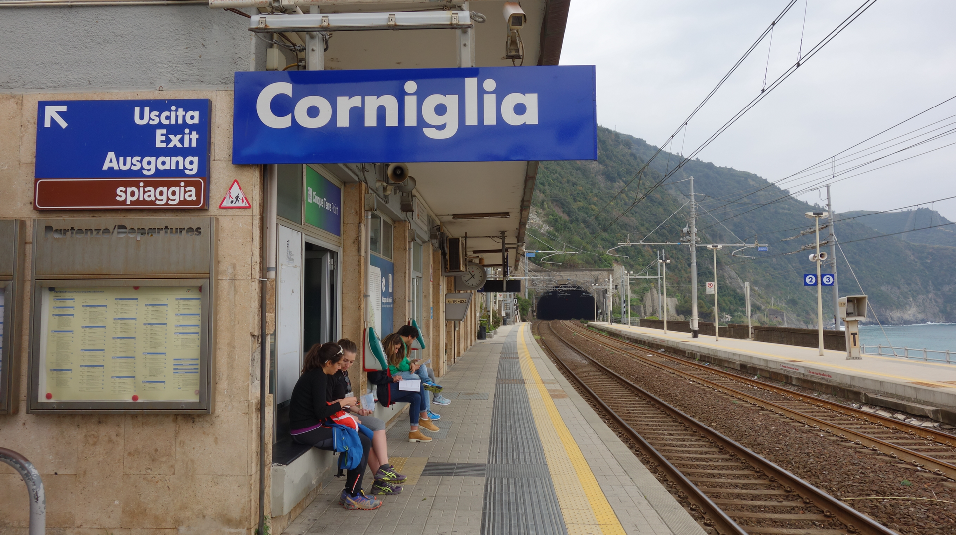 Zdjęcie przedstawia peron na stacji kolejowej we Włoszech. Na tablicy znajduje się nazwa miejscowości: Corniglia. Na peronie znajduje się kilka osób, nie ma pociągu.