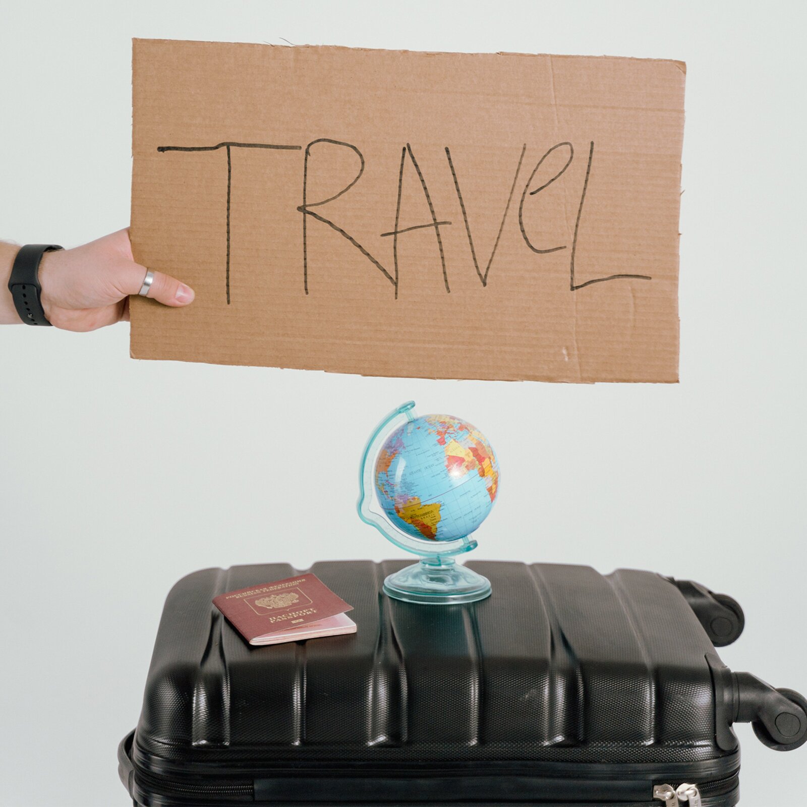 Zdjęcie przedstawia leżącą, czarną, zamkniętą walizkę, na której stoi mały globus i leży paszport. Nad walizką dłoń trzyma kawałek kartonu z napisem "TRAVEL".