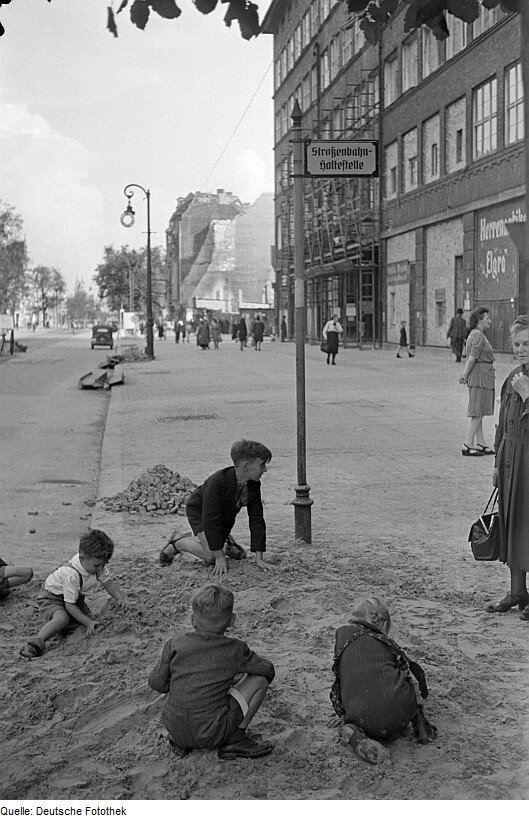 Zdjęcie przedstawia berlińską ulicę w 1945 roku. Na szerokim chodniku, tuż przy ulicy, w piasku bawi się czwórka małych dzieci. Wzdłuż chodnika widać stojące kamienice oraz ruiny zbombardowanych budynków.