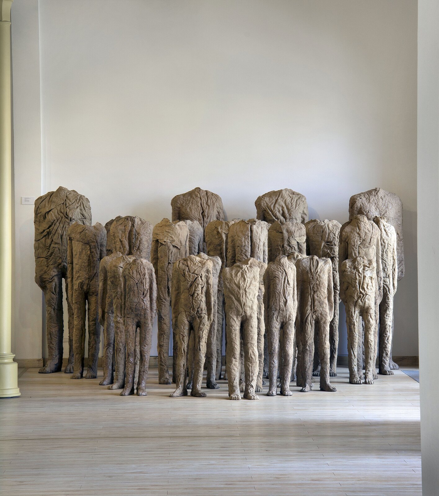 Magdalena Abakanowicz, bez tytułu Fotografia przedstawia rzeźbę znajdującą się w pomieszczeniu o drewnianym parkiecie oraz jasnych ścianach. Z lewej strony znajduje się kremowa kolumna. Rzeźba ukazuje sylwetki kilkunastu postaci, ukazane bardzo blisko siebie, stojąc prosto z opuszczonymi wzdłuż ciała rękami. Struktura postaci jest niejednolita, z licznymi pęknięciami oraz ciemniejszymi elementami na kremowo‑szarej podstawie. Sylwetki pozbawione są głów i szyi, niektóre z nich są wysokie, a inne – niższe, przypominające dziecięce. Umieszczono je w trzech rzędach, od najwyższych do najniższych.