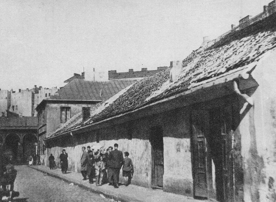 Ilustracja przedstawia czarno‑białą fotografię ulicy z niską, parterową zabudową o zniszczonym, spadzistym dachu. Budynki mają tynki w złym stanie technicznym. Na pierwszym planie widoczna jest grupa osób – głównie mężczyzn i dzieci – stojących i rozmawiających przy ścianie budynku. W tle widać wyższe, wielopiętrowe kamienice. Ulica jest wąska, brukowana lub ubita, bez widocznych elementów infrastruktury miejskiej. 