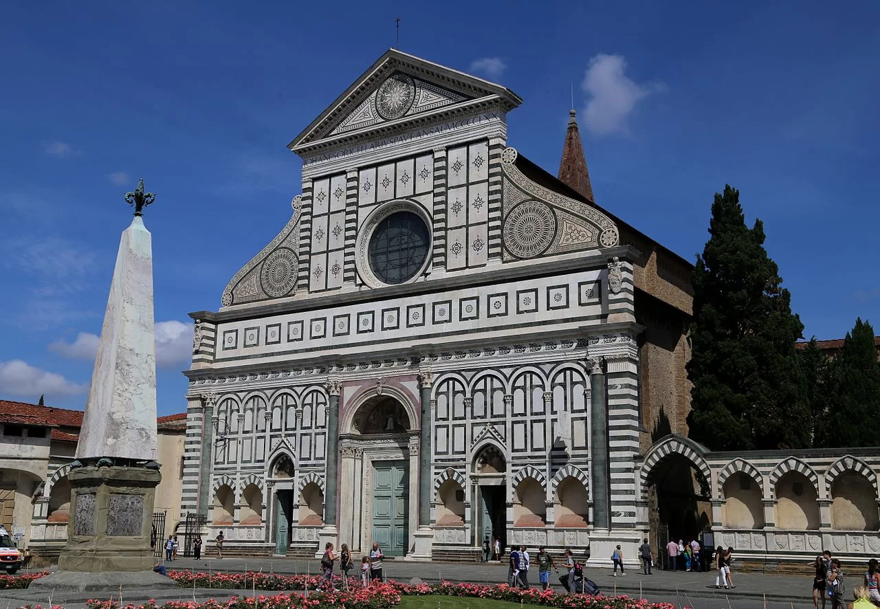 Fotografia przestawia front kościoła Santa Maria Novella we Florencji. Zielone, białe i różowe marmurowe płyty dzielą elewację na mniejsze pola. Górną część, zwieńczoną tympanonem, ograniczają dwie duże woluty, które w łagodny sposób łączą wysoki front nawy głównej ze znacznie niższymi nawami bocznymi. Pośrodku górnej części elewacji umieszczono duże okno – tzw. rozetę, poniżej znajduje się duży portal ozdobiony herbami rodziny Rucellaich, mecenasów kościoła. Do kościoła prowadzi troje drzwi. Po prawej stronie, do budynku kościoła przylega murowane ogrodzenie ozdobione biało‑zieloną arkadą, za którym rosną wysokie drzewa. Po lewej stronie stronie stoi obelisk w kształcie stożka, zakończony lilią. Przed kościołem klomb kwiatowy.