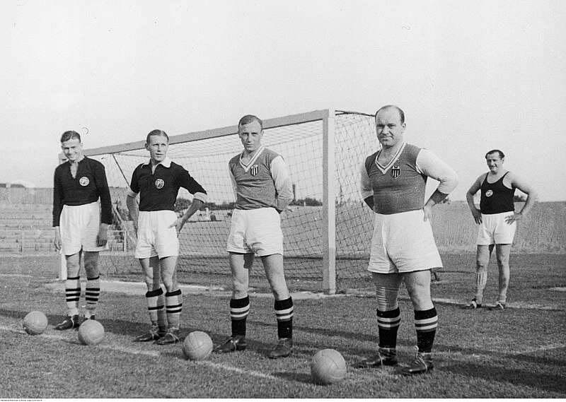 Zdjęcie przedstawia czterech mężczyzn w strojach sportowych. Stoją przed bramką do piłki nożnej. Pod nogami mają piłki.