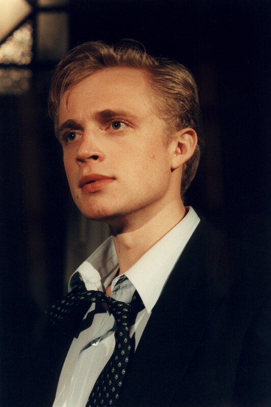 Zdjęcie ze spektaklu pt. "Tango". Na zdjęciu widać twarz młodego mężczyzny. Głowę ma lekko przechyloną w prawo, usta lekko rozchylone. Ma krótkie blond włosy zaczesane na prawą stronę. Ubrany jest w czarną marynarkę i białą koszulę, na której odznacza się czarny krawat w białe wzory.