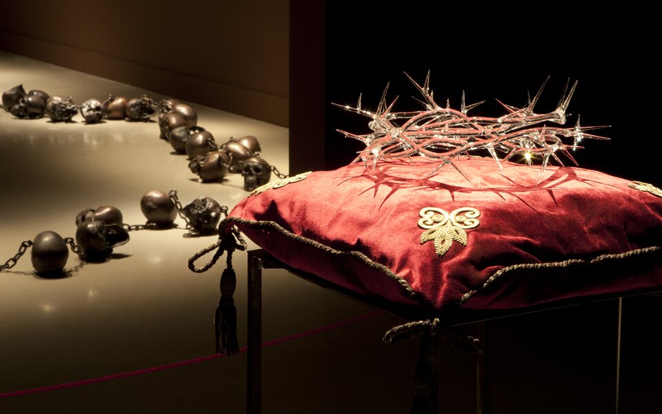 Zdjęcie przedstawia czerwoną, aksamitną poduszkę, bogato zdobioną złotymi nićmi, na której leży korona cierniowa wykonana ze szkła. Poduszka ma w rogach wyhaftowane złote lilie. Jej brzegi obszyte są złotym sznurem zawiązanym na rogach w pętelki. Po lewej stronie, na drugim planie widać metalowe czaszki, leżące w nierównym rzędzie na podświetlonej podłodze. Są one powiązane łańcuchem. Tło jest czarne.