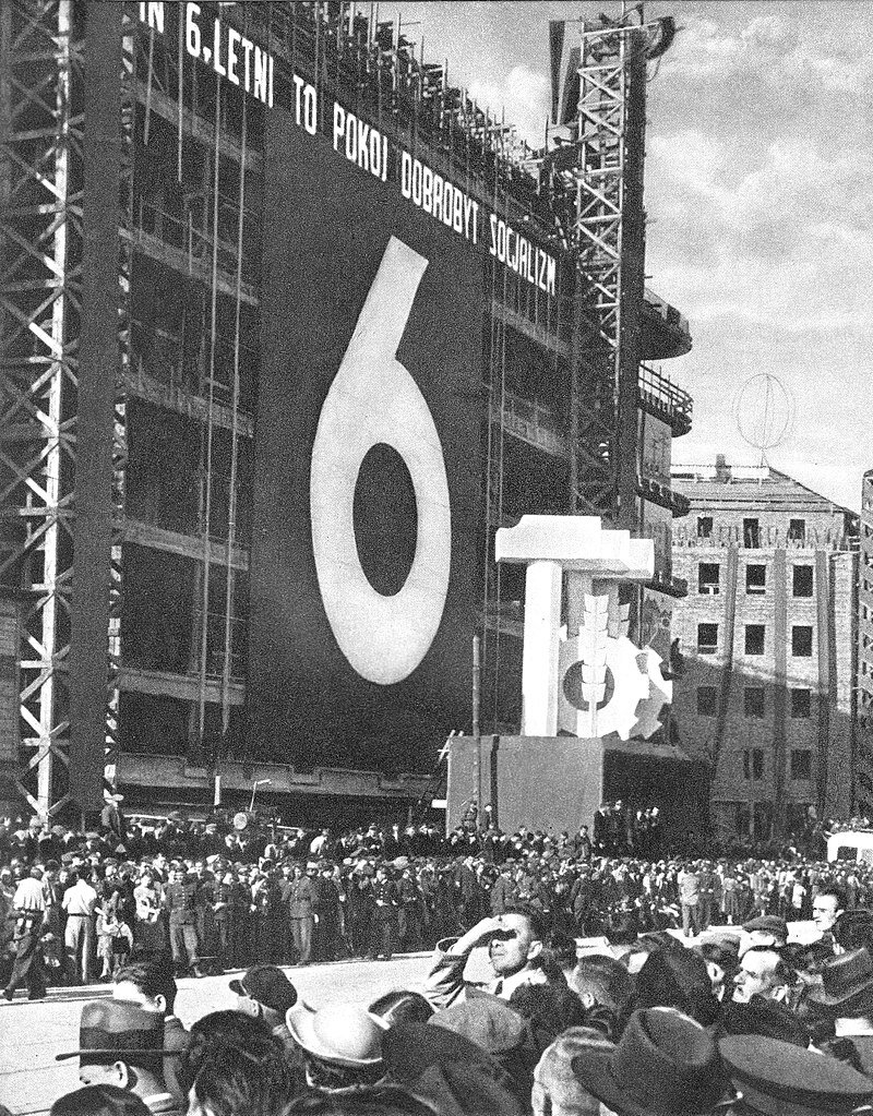 Fotografia przedstawia tłum ludzi stojących pod jednym z nowo budowanych budynków, na którego ścianie wisi wielkie płótno z ogromną cyfrą sześć. Tytuł płótna brzmi: sześcioletni to pokój dobrobyt socjalizm.