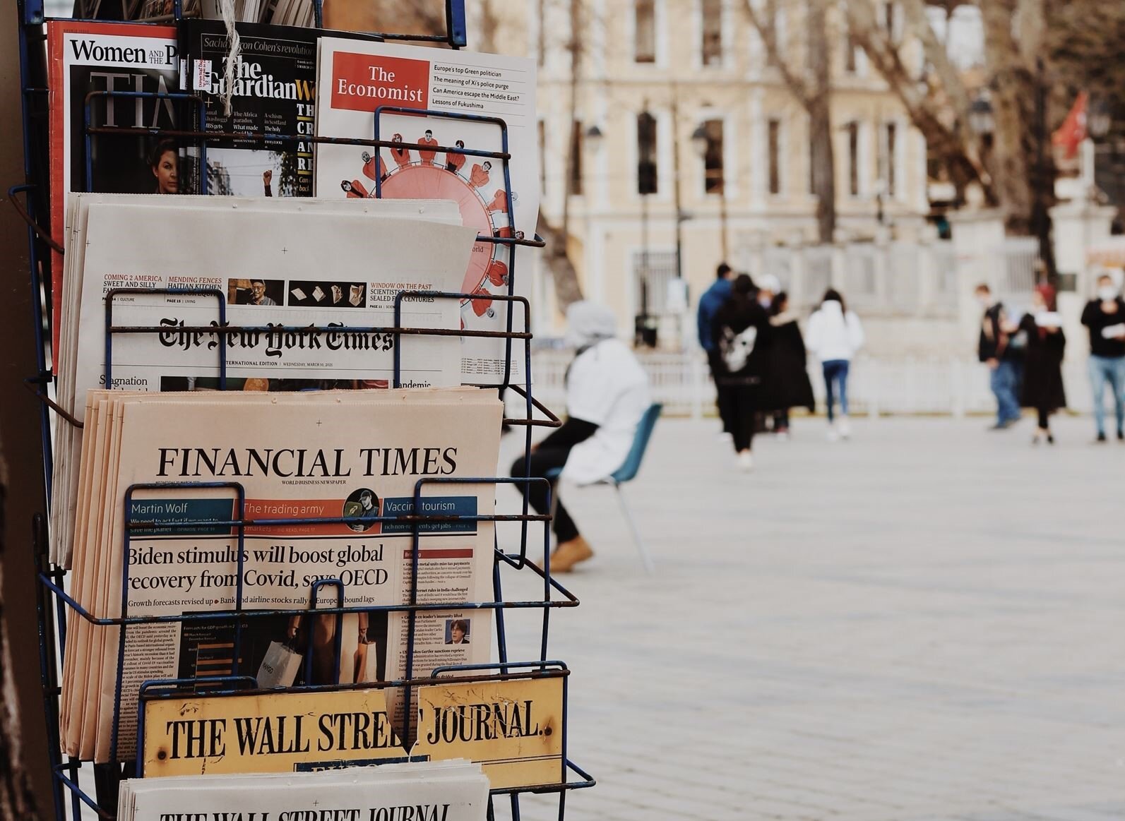 Zdjęcie przedstawia po lewej stronie metalowy stojak, w którym widoczne są poukładane w stosy gazety. Widoczne są tytuły gazet w języku angielskim: FINANCIAL TIMES, THE WALL STREET JOURNAL, THE GUARDIAN, THE NEW YORK TIMES. Pośrodku i po prawej stronie widoczny jest plac, zaś w tle fasady budynków. Na placu widoczne są przechodzące osoby.