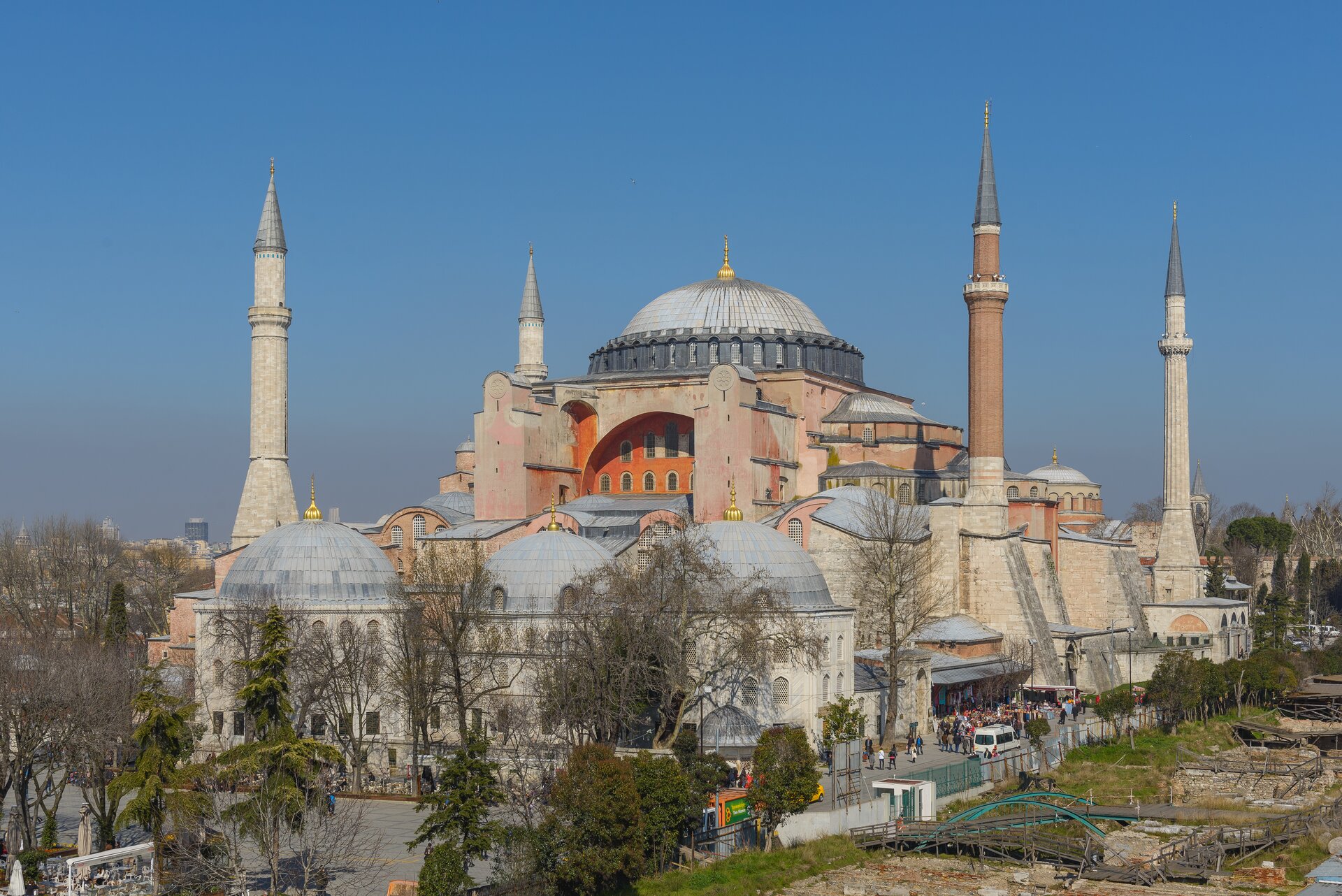 Na zdjęciu znajduje się Hagia Sophia. To rozbudowana bazylika z dużą kopułą w centralnym miejscu i czterema wysokimi minaretami – wąskimi wieżyczkami w każdym z jej rogów. Świątynia ma także liczne przedsionki z wejściami, przed jednym z nich – po prawej stronie zdjęcia są czekający na wejście turyści. Budowla zbudowana na planie centralnym utrzymana jest w kolorze piaskowym, ceglastym, pomarańczowym i szarym. Dachy kopuł pokryte zostały srebrno‑szarymi dachówkami, na samych szczytach wykończonymi złotymi elementami. Przed bazyliką znajduje się plac, przy którym rosną drzewa. Niebo jest bezchmurne.