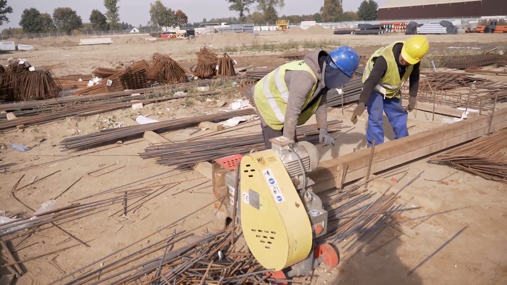 Zdjęcie przedstawia dwóch robotników ubranych w kamizelki odblaskowe i kaski, obsługujących maszynę do cięcia stali.