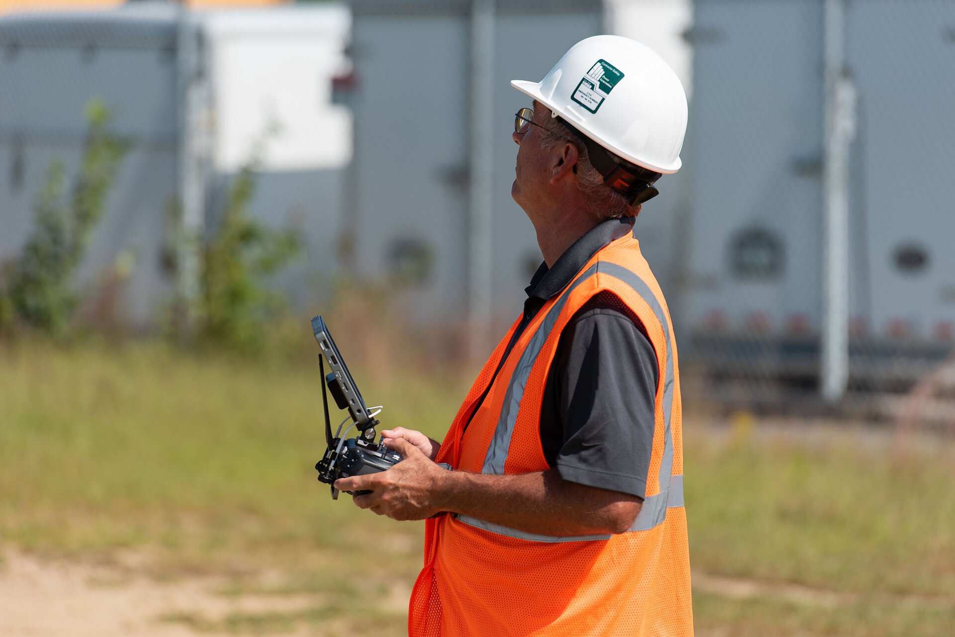 Zdjęcie przedstawia mężczyznę w kasku ochronnym oraz odblaskowej kamizelce, trzymającego w rękach joystick z ekranem. Mężczyzna patrzy w górę. 