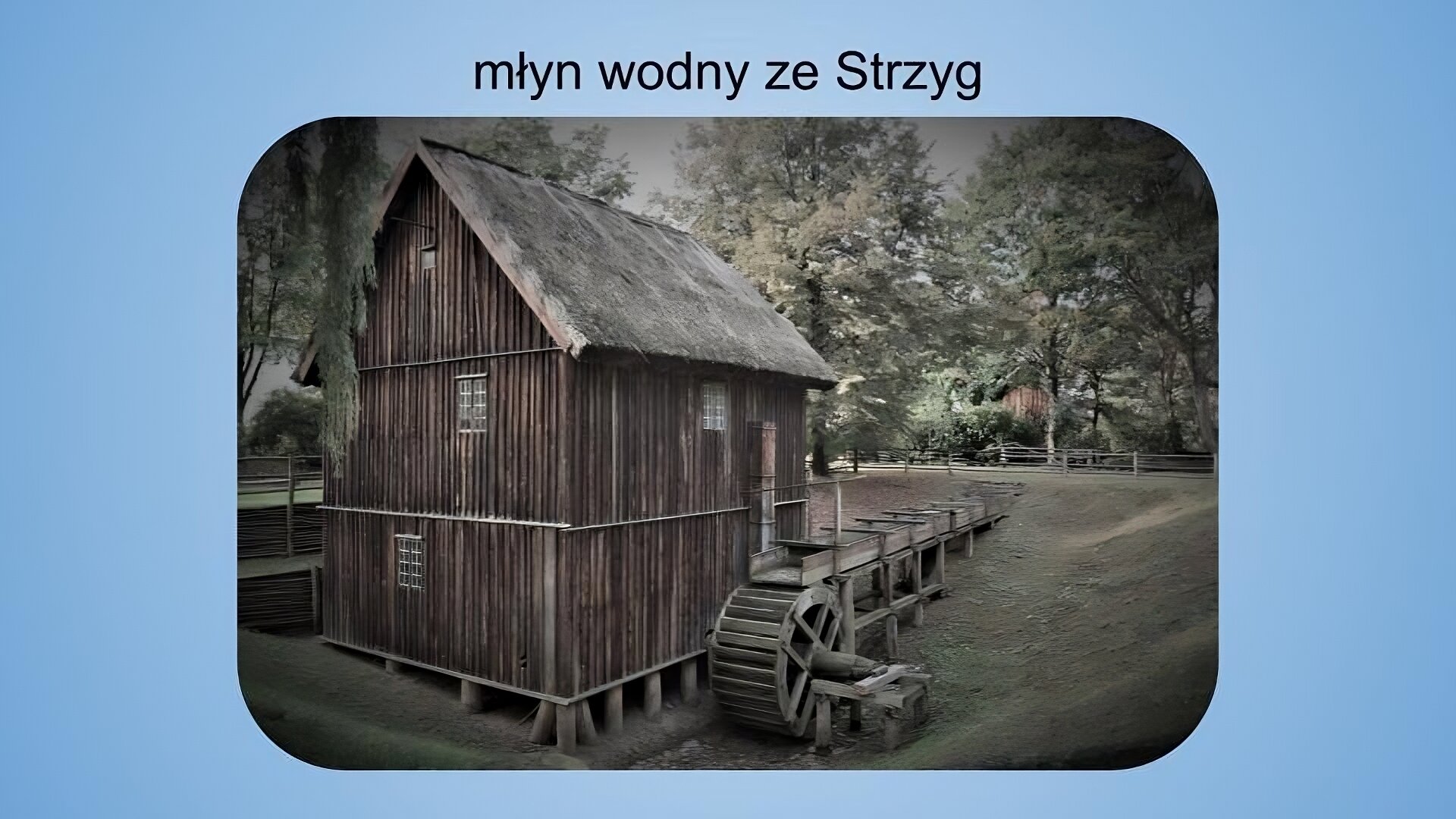 Jasnoniebieskie tło. Na górze napis: młyn wodny ze Strzyg. Poniżej kolorowa fotografia przedstawiająca drewnianą konstrukcję młyna ujętą od tyłu. Widoczna jest tylna ściana budynku z dwoma średniej wielkości oknami i jesnym niewielkim oknem na poddaszy oraz jedna z bocznych ścian budynku z drzwiami wejściowymi i jednym oknem. Obok ściany bocznej znajduje się koło młyńskie. Na dalszym planie widać pomost prowadzący do drzwi frontowych. Dach budynku jest spadzisty i pokryty strzechą. W tle widać drzewa iglaste, a na ziemi równo przystrzyżoną trawę. Zdjęcie jest nieco przyciemnione.