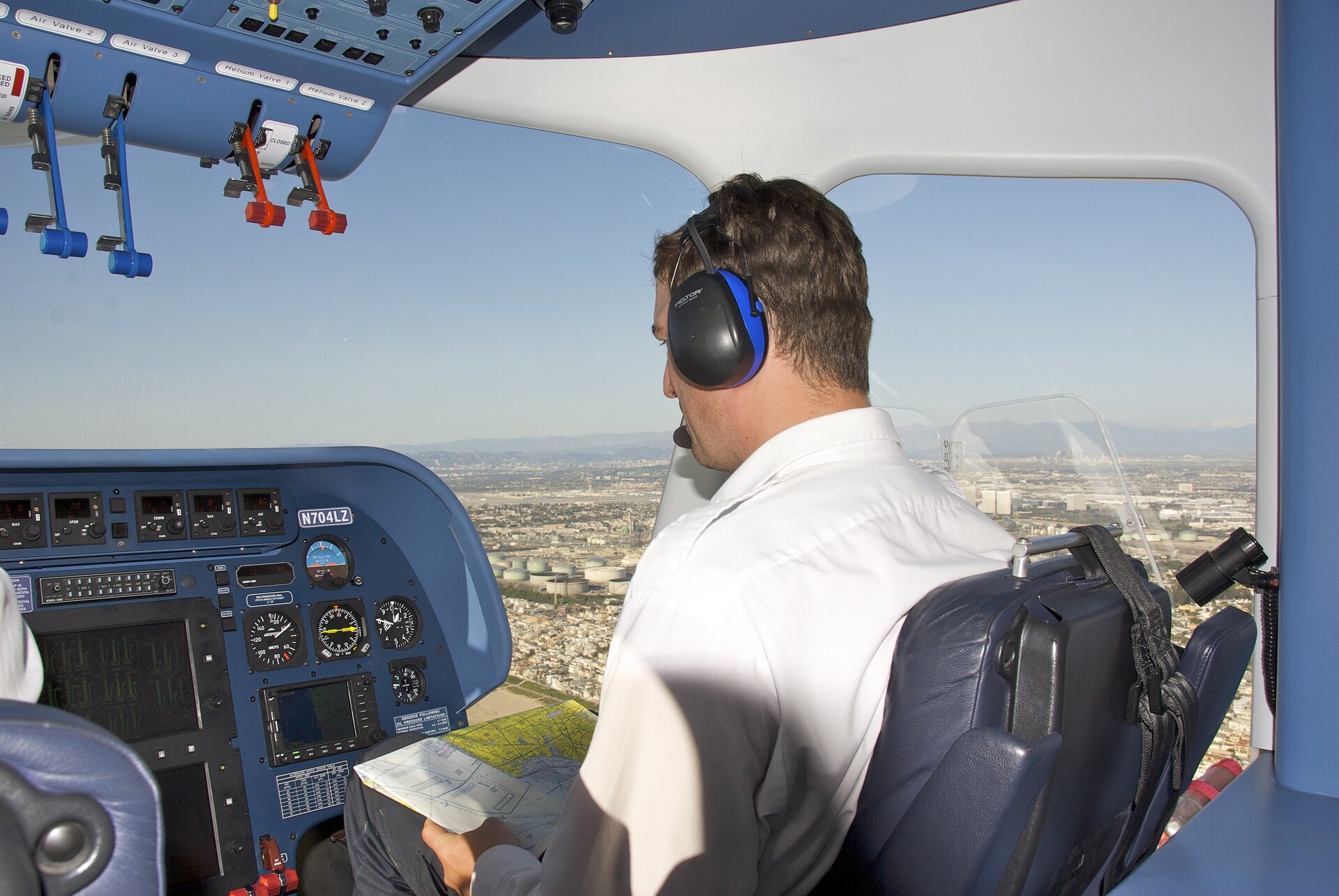 Ilustracja przedstawiająca pilota siedzącego w kokpicie tyłem do kadru. Pilot ma na głowie słuchawki z mikrofonem, na kolanach trzyma mapę. 
