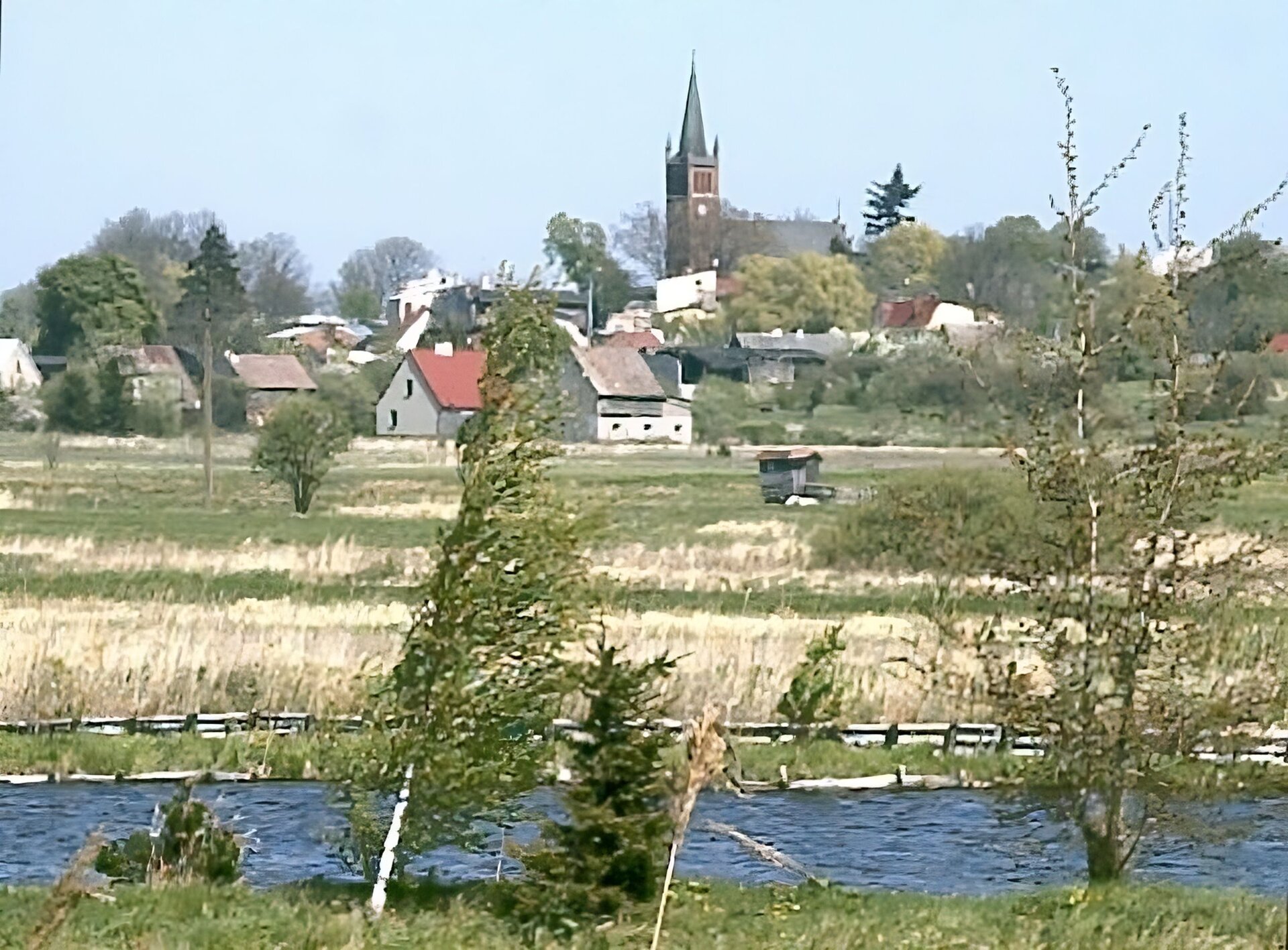 Na fotografii na pierwszym planie znajduje się kilka drzew. Za nimi jest rzeka, a następnie polana. Dalej znajdują się zabudowania. Nad domami góruje wieża kościoła. W tle niebo.