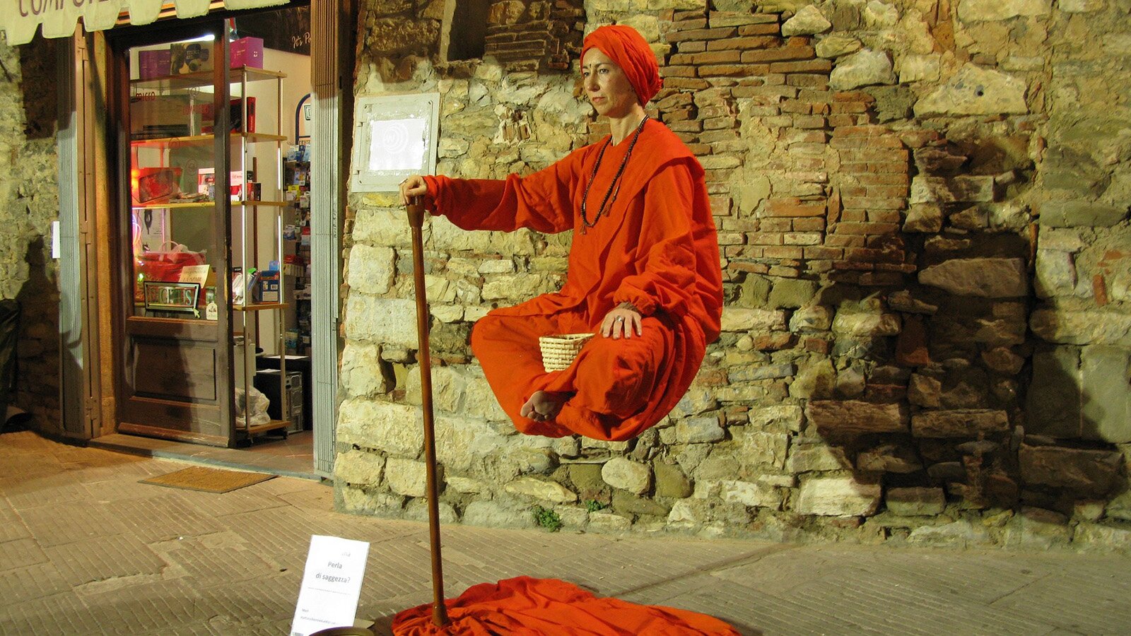 Zdjęcie przedstawia osobę w turbanie i luźnym stroju, lewitującą w powietrzu, jedyne na czym się opiera jedną ręką to kij.