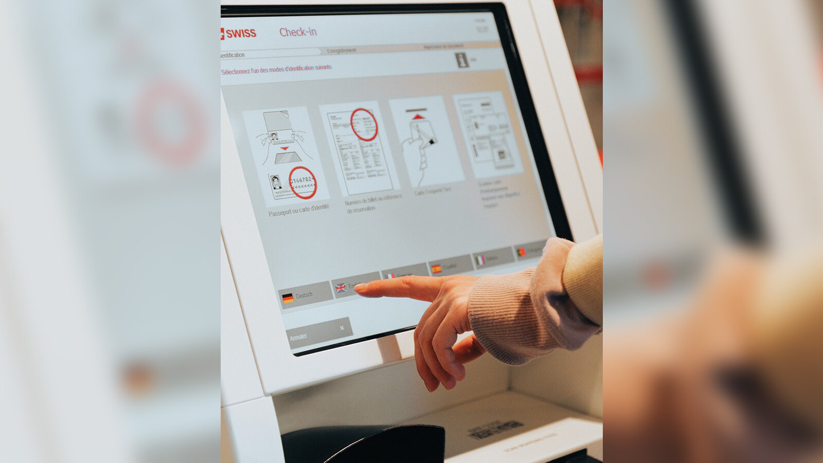 Zdjęcie przedstawia około 20 calowy monitor na górze którego ukazano napis "Swiss check-in". Przy dolnej części ekranu jest dłoń z wysuniętym palcem wskazującym.