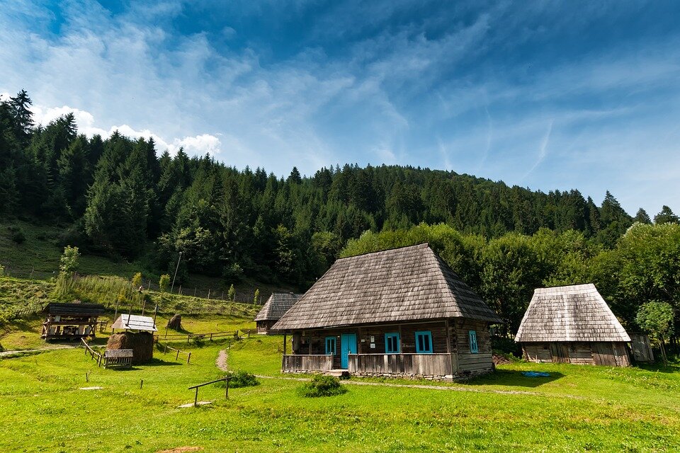 Zdjęcie przedstawia Karpaty. Na pierwszym planie jest trawiasty teren. Stoją na nim drewniane chaty o stromych dachach. Jeden z nich ma niebieskie drzwi i okiennice. Pomiędzy zabudowaniami biegnie ścieżka. Za chatami rośnie las iglasty.