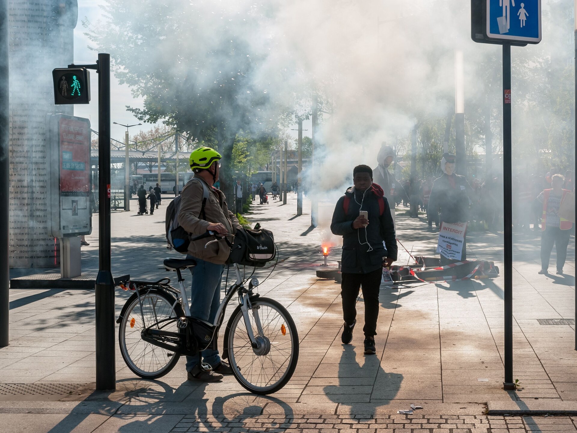 Zdjęcie przedstawia plac na którym jest silne zadymienie, ponieważ odbywa się demonstracja, palone są race, prowadzona jest transmisja telewizyjna której przygłąda się mężczyzna na rowerze w zielonym kasku.