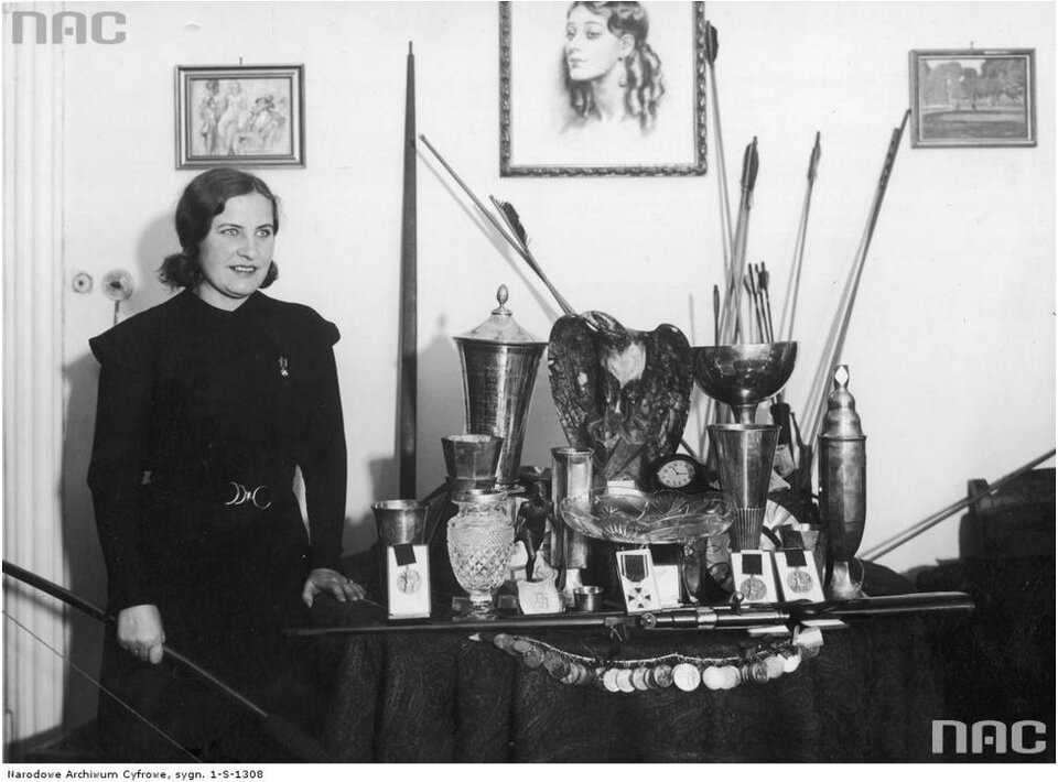 Zdjęcie przedstawia kobietę w ciemnej sukni, która stoi przy stoliku. Na stoliku są trofea i medale.
