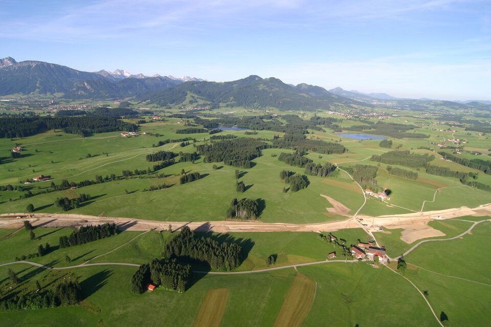 Zdjęcie przedstawia płaski teren porośnięty trawą i drzewami, a w tle pasmo górskie. Na płaskim terenie odbywa się budowa drogi.  