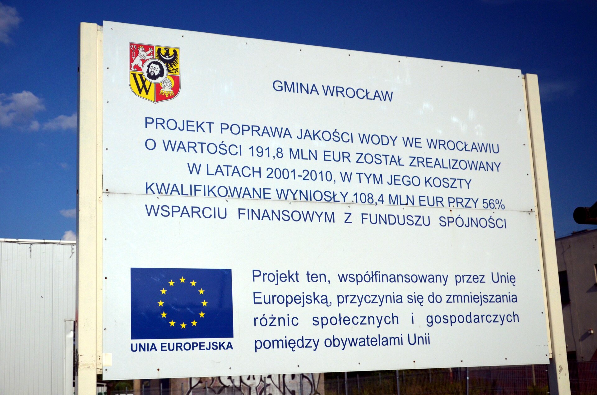 Na zdjęciu biała tablica z herbem Wrocławia i flagą Unii Europejskiej z napisem: Gmina Wrocław, projekt poprawa jakości wody we Wrocławiu o wartości sto dziewięćdziesiąt jeden i osiem dziesiątych miliona euro został zrealizowany w latach dwa tysiące jeden – dwa tysiące dziesięć, w tym jego koszty kwalifikowane wyniosły sto osiem i cztery dziesiąte miliona euro przy pięćdziesięcio sześcio procentowym wsparciu finansowym z funduszu spójności. Projekt ten, współfinansowany przez przez Unię Europejską, przyczynia się do zmniejszania różnic społecznych i gospodarczych pomiędzy obywatelami Unii.