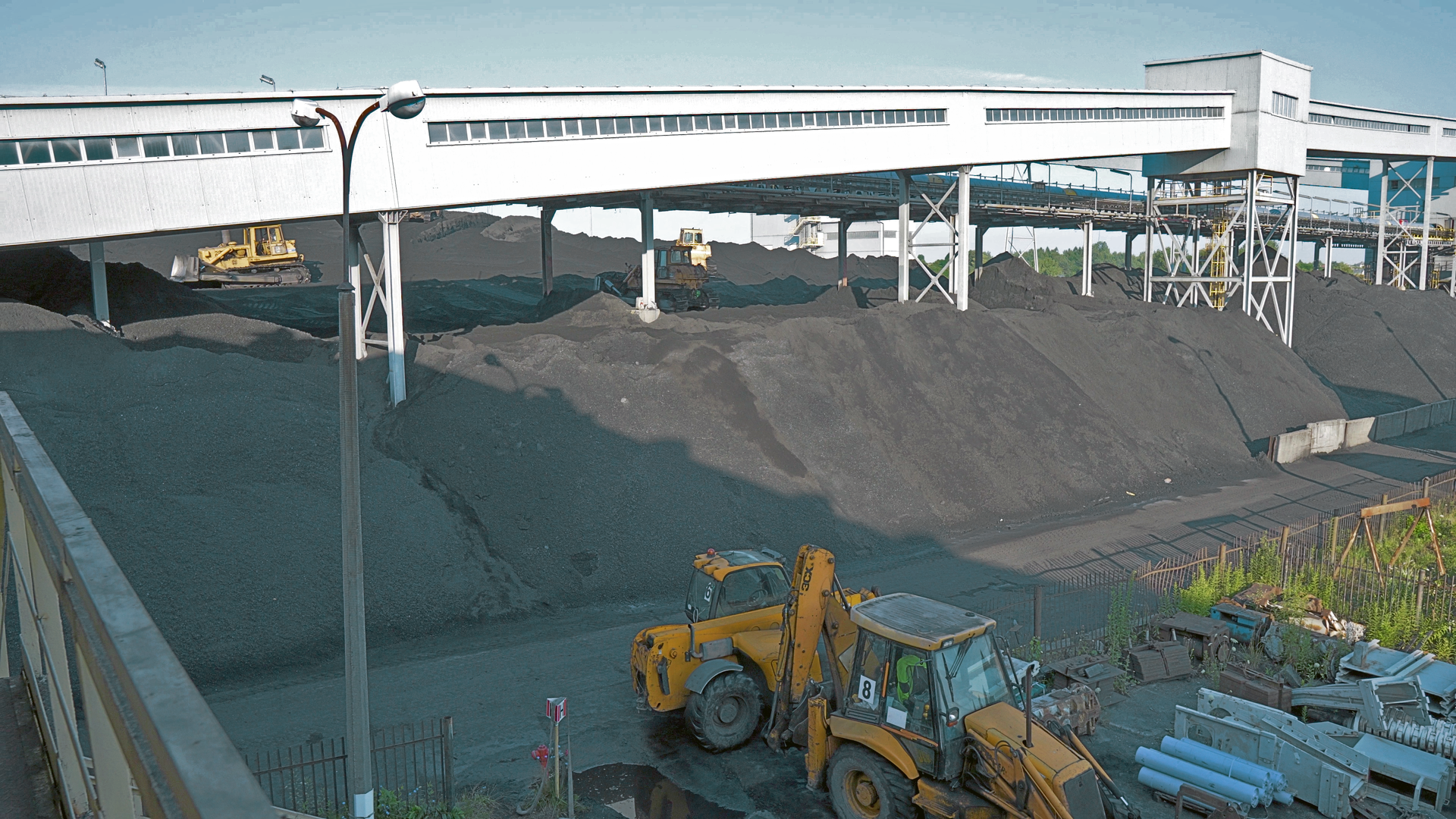 Na zdjęciu widoczne jest składowisko z węglem. Widoczne na zdjęciu zwały mają kształt pryzm. Przed pryzmami znajdują się dwie żółte koparki. W tle widać zabudowany przenośnik taśmowy. Przenośnik jest podwieszony. Przenośnik zapada stronę gruntu.