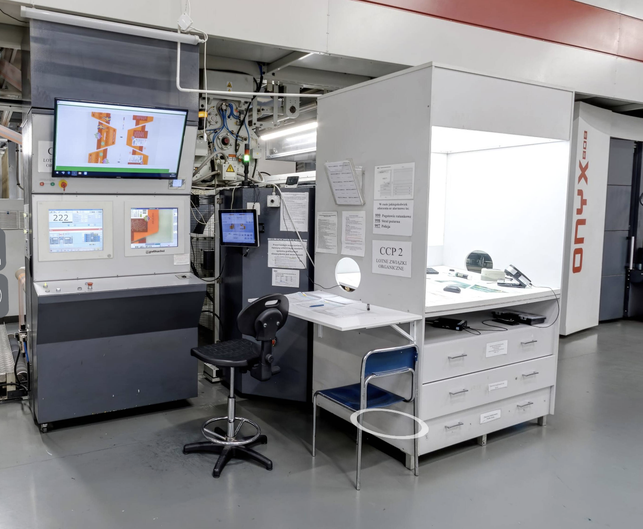 Zdjęcie przedstawia przykładowe miejsce kontroli jakości druku w drukarni. Po lewej widoczne jest stanowisko z monitorami, po prawej stanowisko do wykonywania kontroli za pomocą urządzeń, np. densytometru, spektrometru. Widoczna jest szafka z podświetlonym wgłębieniem, w której na blacie ułożone są różne przyrządy. Na boku szafki widoczny jest wystający prostokątny blat, do którego dostawione jest niebieskie krzesło. Pomiędzy stanowiskiem z monitorami a stanowiskiem kontroli widoczne jest czarne obrotowe krzesło. W tle widoczne są maszyny poligraficznej. 