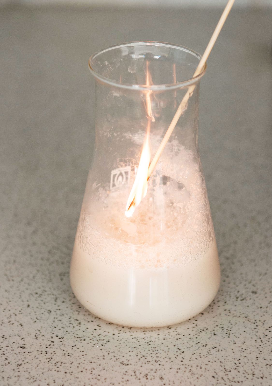 Zmienić opis alt na: Zdjęcie kolby, do której włożone jest zapalone łuczywo. W kolbie biała pieniąca się mieszanina drożdży i wodnego roztworu nadtlenku wodoru.