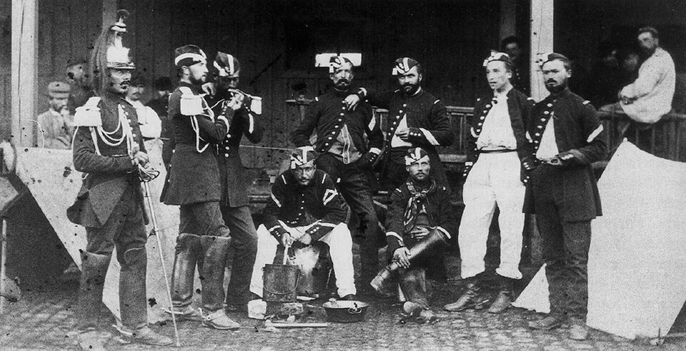 Zdjęcie przedstawia grupę mężczyzn w mundurach. Dwóch mężczyzn siedzi przed budynkiem, a kilku stoi w półkolu za nimi. Żołnierze mają na sobie mundury i czapki z materiału.  Po lewej stronie stoi żołnierz w wysokim hełmie na głowie. W dłoniach trzyma szablę. 