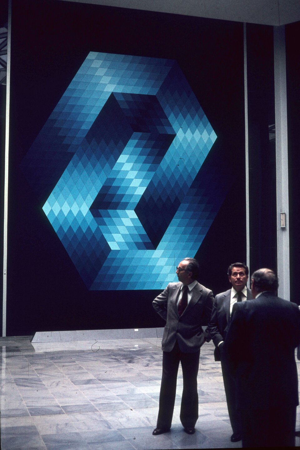 Ilustracja przedstawia Victora Vasarely w towarzystwie dwóch innych mężczyzn w pomieszczeniu w siedzibie Fundacji Vasarely'ego. Za mężczyznami, na granatowej ścianie widoczne jest duże dzieło. Jest to figura złożona z małych rombów w różnych odcieniach koloru niebieskiego.