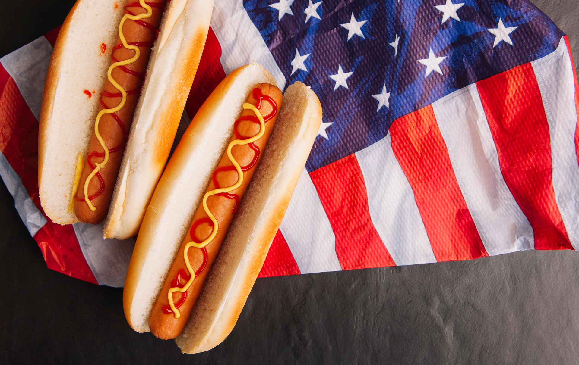 Zdjęcie przedstawia widoczne z góry dwa hot-dogi po lewej stronie zdjęcia, które leżą na zagiętej amerykańskiej fladze. Całość znajduje się na ciemnoszarym tle imitującym fakturę papieru. Hot-dogi są lekko przyrumienione, a parówki w środku polane wężykami keczupu i musztardy.