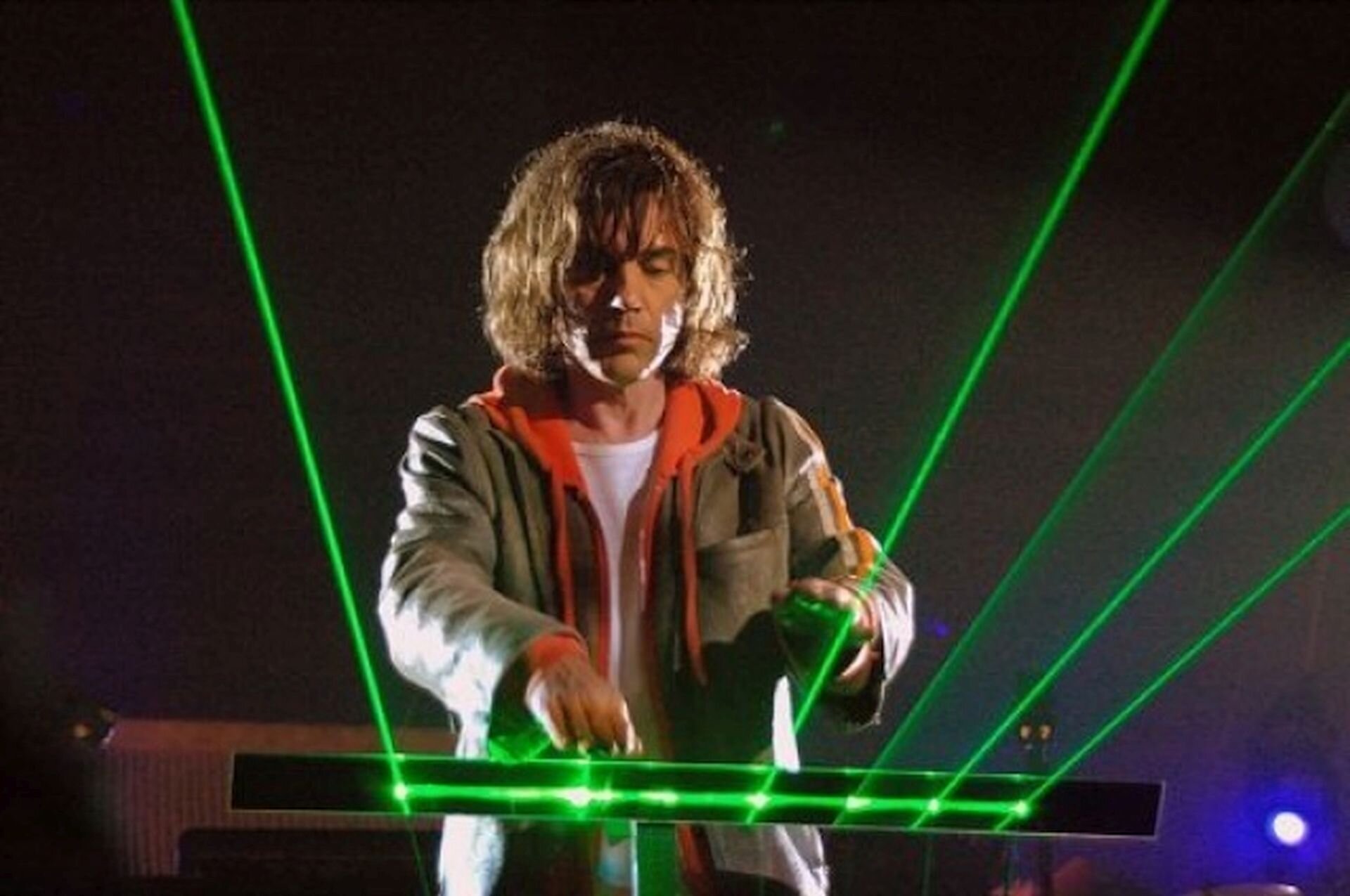 Na zdjęciu Jean‑Michel Jarre, grający na harfie laserowej. Mężczyzna ma włosy do ramion. Ubrany jest w białą koszulkę, czerwoną bluzę z kapturem oraz szarą marynarkę. Gra na instrumencie, z którego wydobywają się zielone wiązki laserowe.