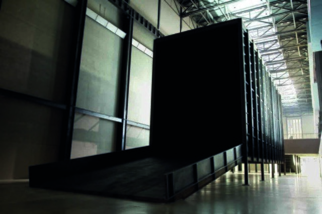 Fotografia przedstawia halę z wysokim dachem, w której umieszczony został czarny kontener stojący wzdłuż lewej ściany hali. Jest to instalacja Mirosława Bałki pod tytułem „Jak to jest?”. Kontener zbudowany jest ze stalowych ścian, a stoi na metalowych palach. Prowadzi do niego opuszczona na szarą podłogę rampa. Ściana hali również jest szara, przebiegają przez nią metalowe elementy. U góry, przez siatkę metalowych konstrukcji, można zauważyć wpadające przez sufit światło.