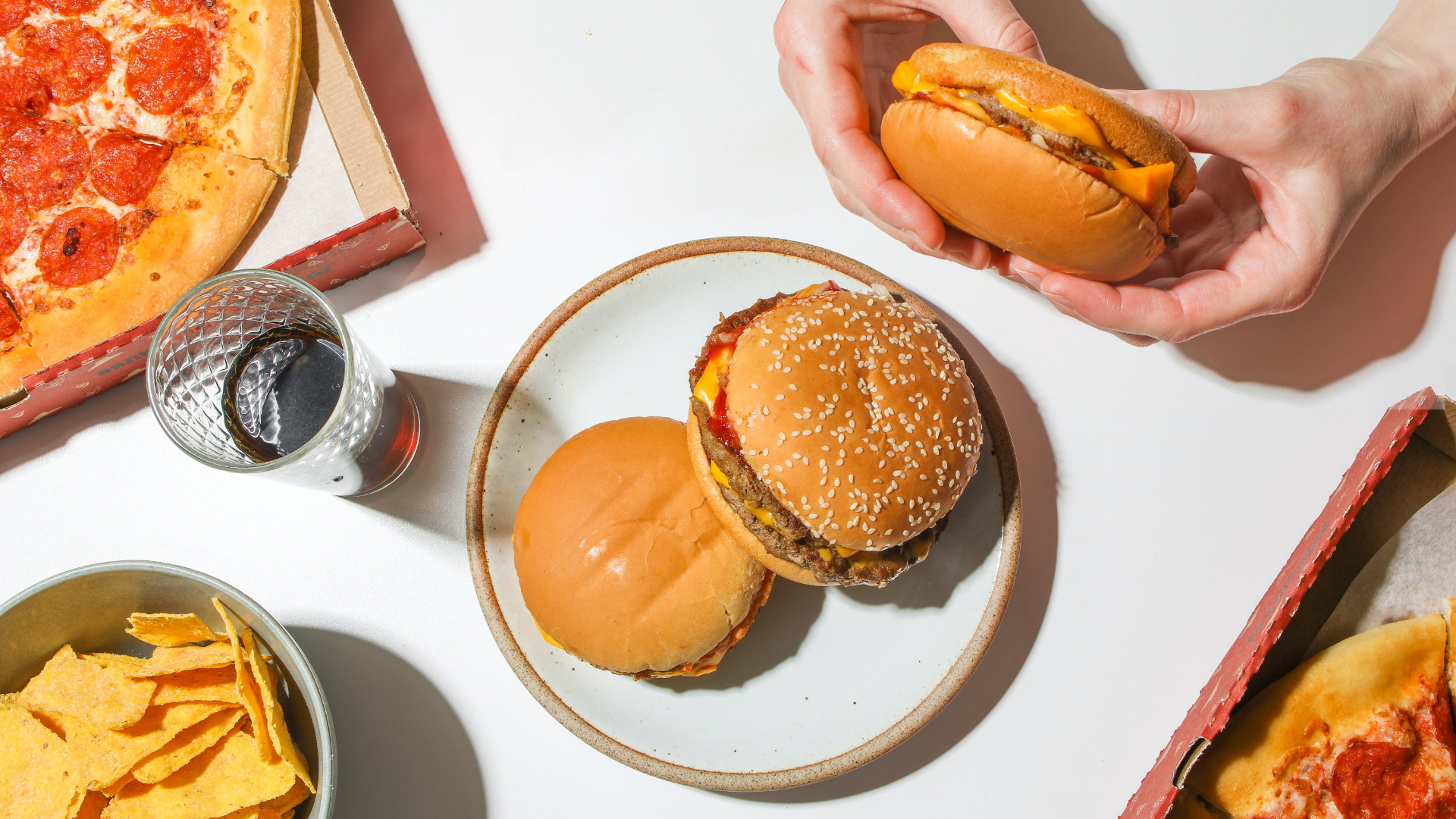 Zdjęcie przedstawia produkty typu fast food. Na środku zdjęcia na talerzu leżą dwa hamburgery. Po prawej stronie, na górze widoczne są ręce osoby, która trzyma trzeciego hamburgera. Po lewej stronie, na górze widoczny jest fragment pizzy z salami w pudełku. Po lewej stronie, na dole stoi miseczka z czipsami. Po między pizzą a miseczka stoi szklanka wypełniona ciemnym napojem.