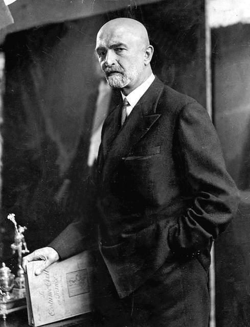 Zdjęcie przestawia mężczyznę w średnim wieku w pozycji stojącej. Ubranego w ciemny garnitur i białą koszulę. W prawej ręce trzyma książkę, którą opiera o stolik. Lewą rękę ma włożoną do kieszeni spodni. Nie ma włosów. Na twarzy wąsy i broda.