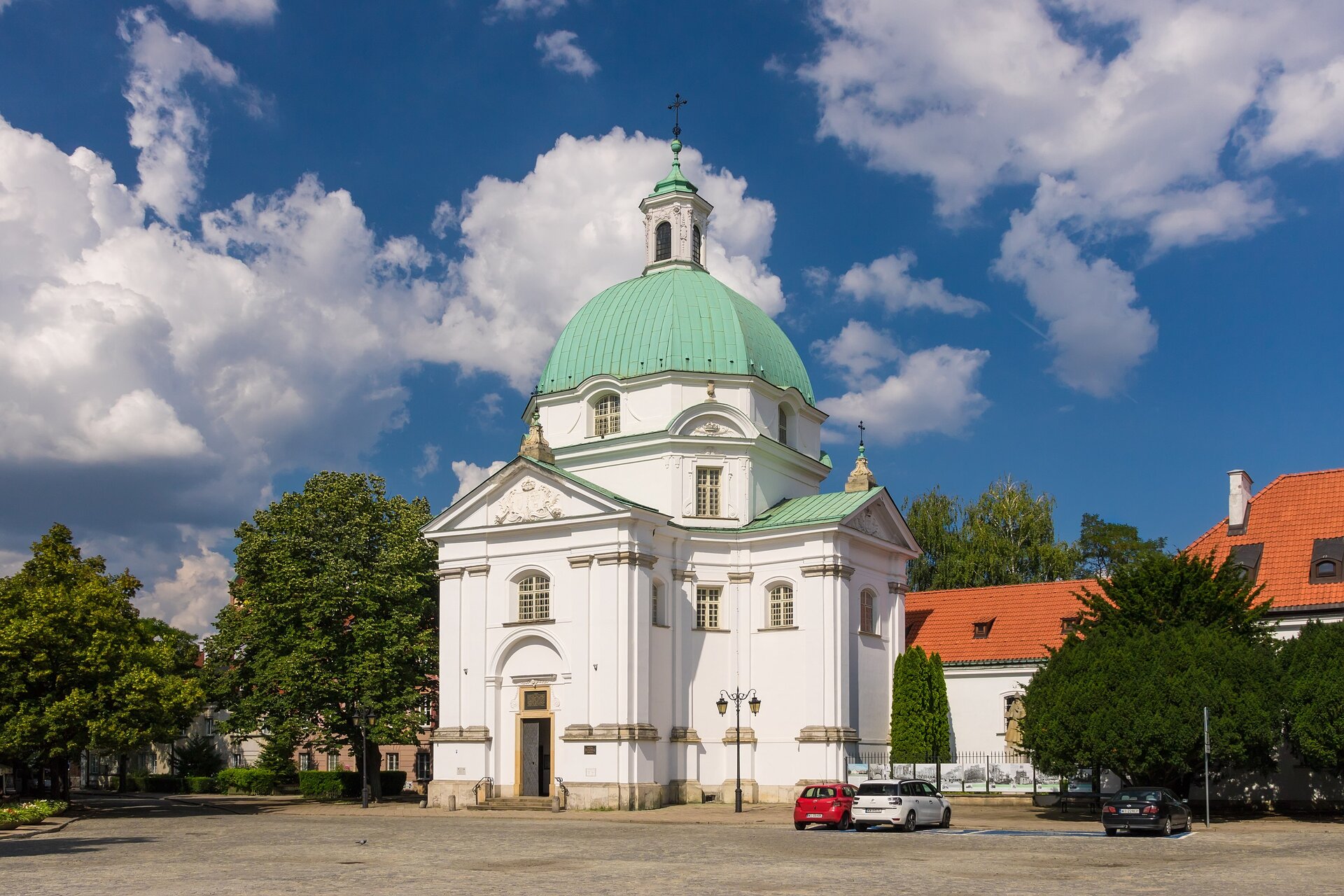 Ilustracja przedstawia kościół św. Kazimierza na Rynku Nowego Miasta, widok współczesny. Jest to mały kościółek o jasnej elewacji. Posiada dużą kopułę o jasnozielonym obiciu z wieżą na czubku. Znajduje się zaraz przy drodze, obok zaparkowane są samochody.