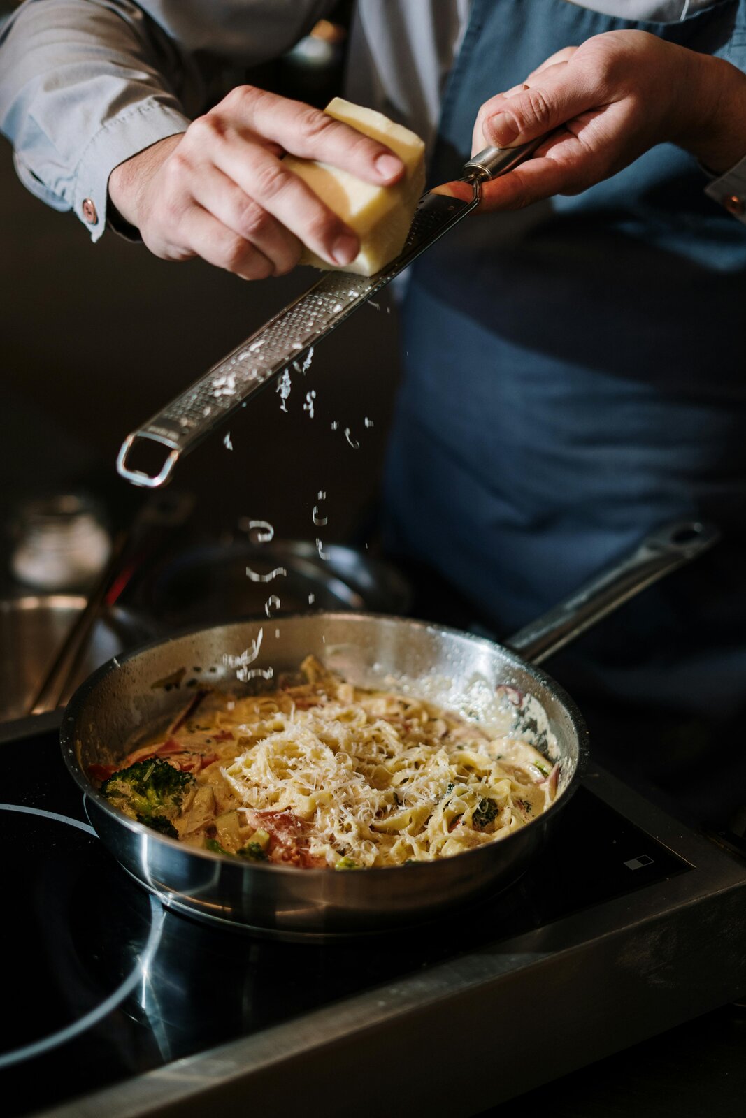 Na zdjęciu widać, jak mężczyzna ubrany w szarą koszulę i niebieski fartuch trze świeży kawałek parmezanu nad patelnią. W patelni znajduje się kremowe danie makaronowe z różnymi dodatkami.