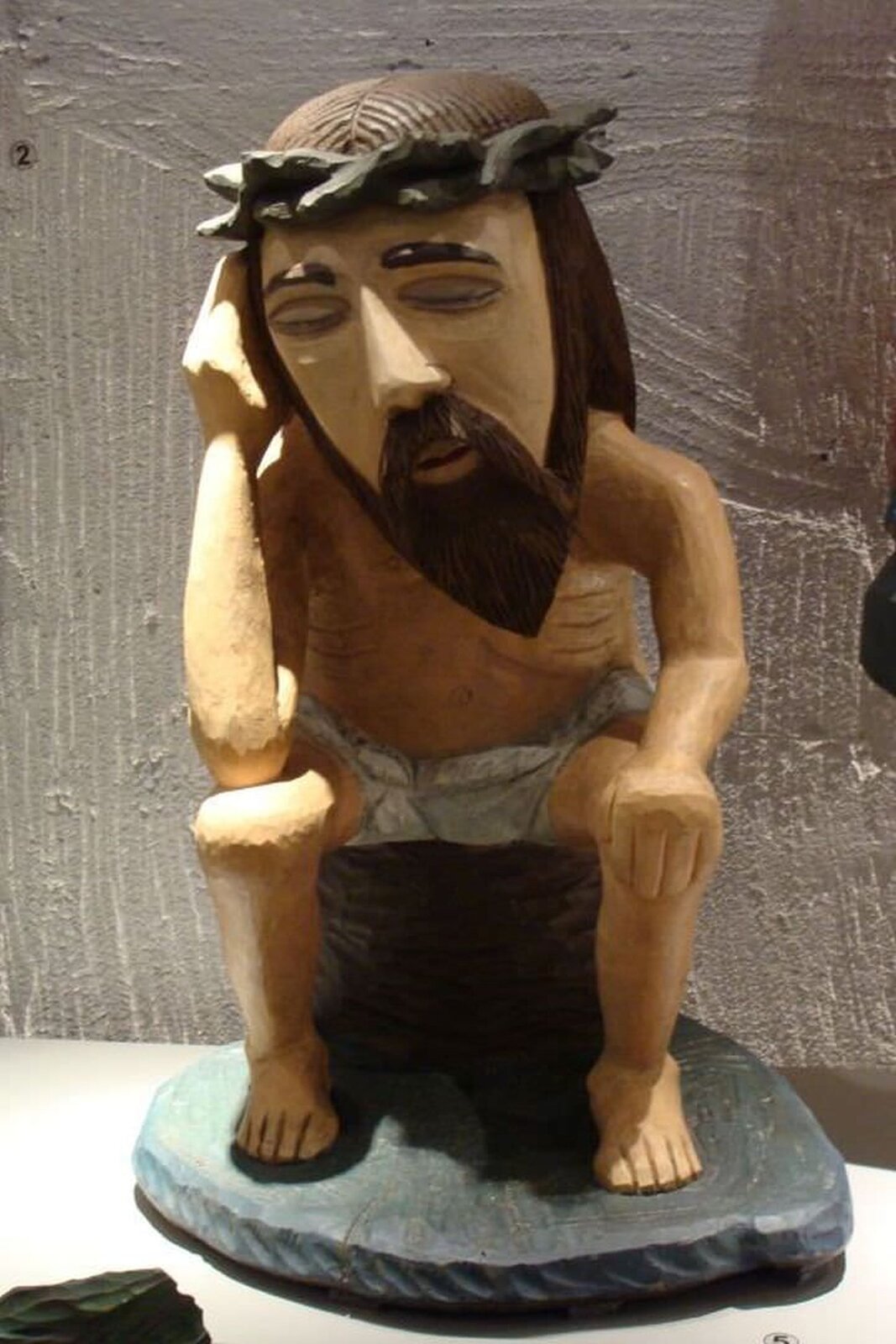 Ilustracja przedstawia drewnianą rzeźbę Konstantego Marcinkowskiego „Chrystus frasobliwy”. Ukazuje Jezusa z zamkniętymi oczami. Na głowie ma koronę cierniową. Jest ukazany w pozycji siedzącej, prawą ręką podpiera głowę.