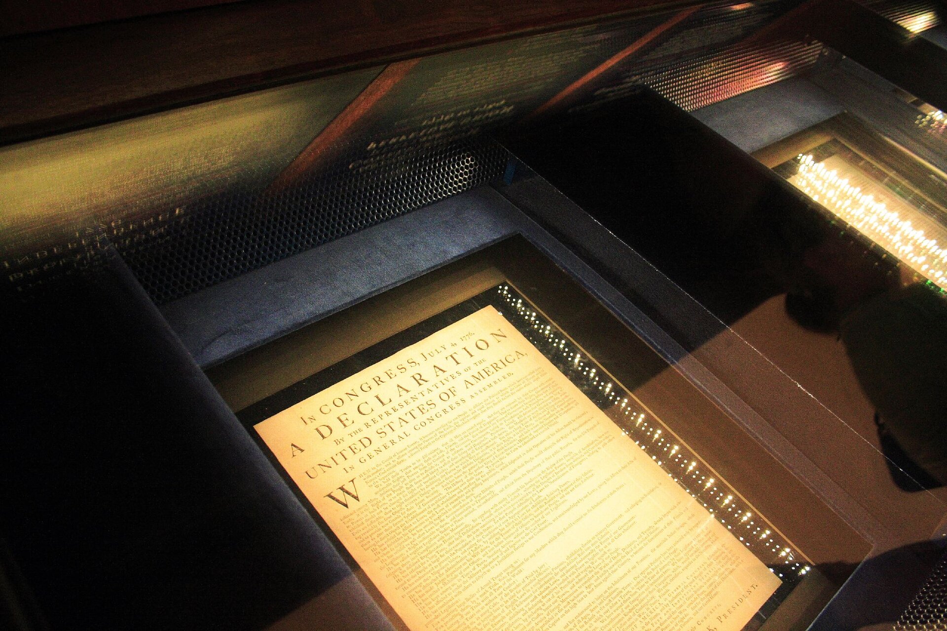 Ilustracja przedstawia zdjęcie Deklaracji Niepodległości Stanów Zjednoczonych przechowywanej w szczelnie zamkniętej oszklonej gablocie. Dokument jest oświetlony otaczającymi go paskami diod LED w celu umożliwienia jego łatwego odczytania przez kilka warstw szkła.