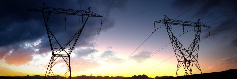 Zdjęcie przedstawia dwa słupy wysokiego napięcia z rozpiętymi przewodami na tle zachodzącego słońca i ciemniejącego nieba.