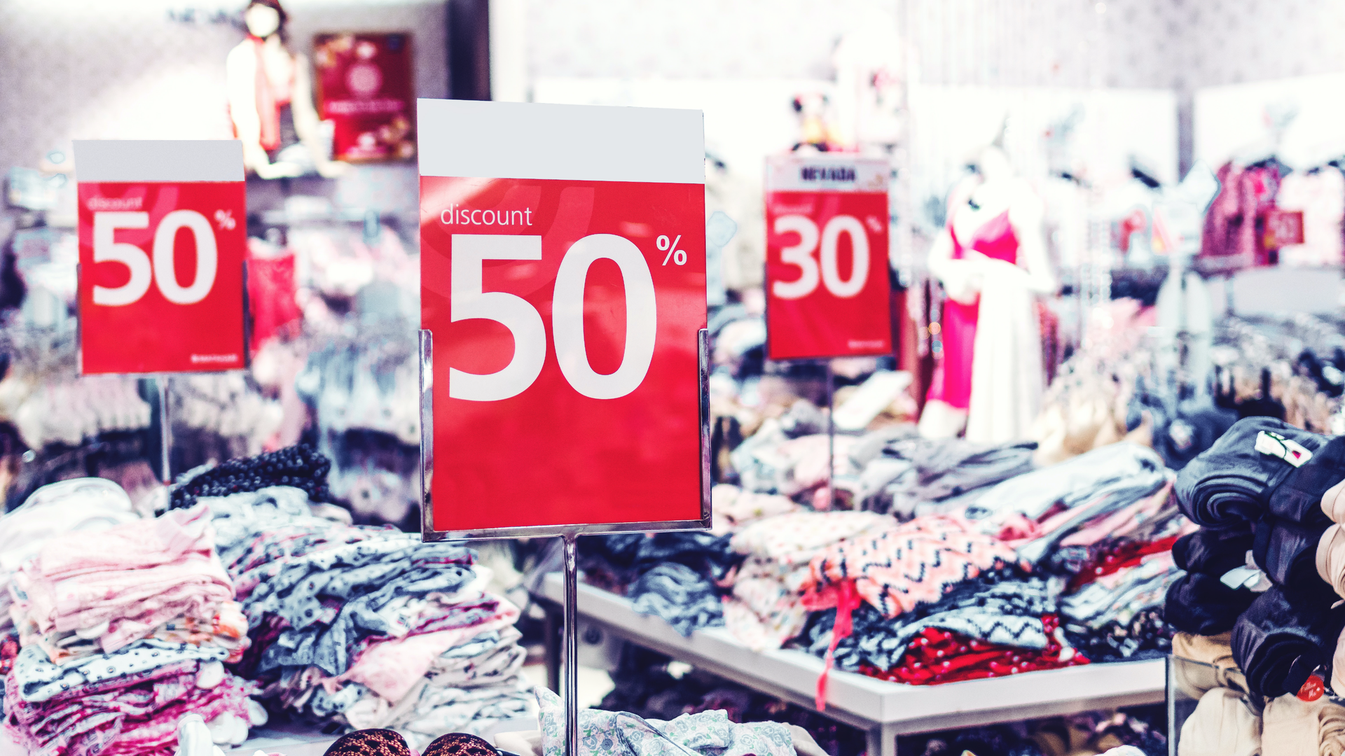 Na zdjęciu znajduje się wnętrze sklepu z dużą ilością ubrań położonych na stołach. Na stołach obok ubrań znajdują się plakaty informujące o przecenie. Dwa z napisem discount 50% oraz jeden z napisem discount 30%.