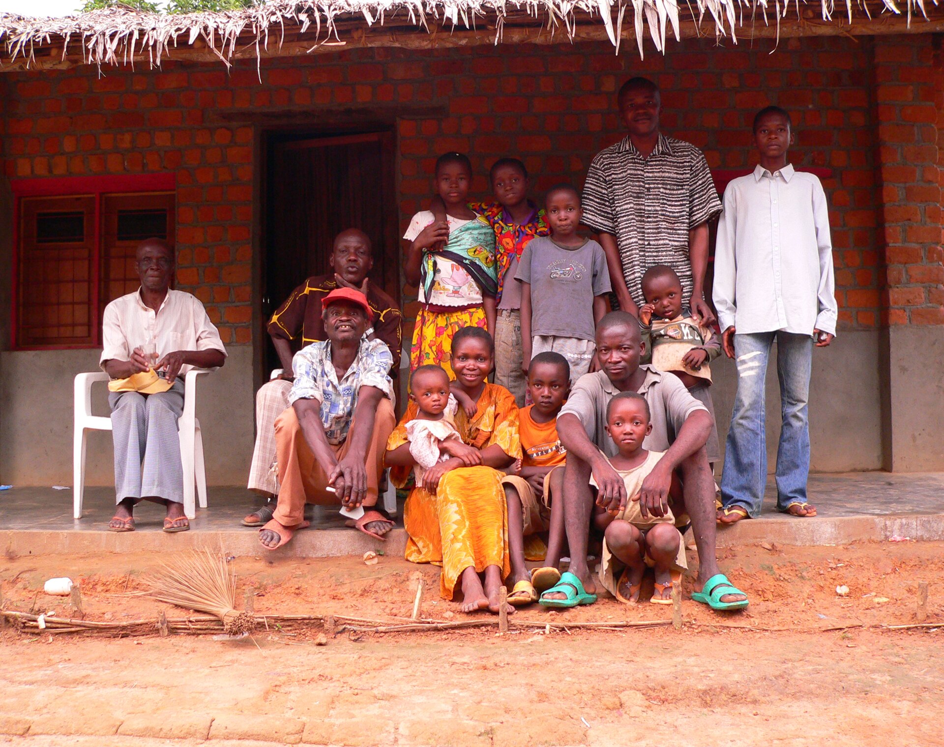 Fotografia przedstawia grupę czarnoskórych ludzi w różnym wieku znajdujących się przed murowanym domem. Stoją w dwóch rzędach: kilka osób siedzi, pozostali stoją. Ubrani są w kolorowe ubrania. Wszyscy mają krótko ostrzyżone włosy. Część się uśmiecha. Po lewej stronie, w niewielkim oddaleniu siedzi na krześle starszy mężczyzna z czapką z daszkiem ułożoną na kolanach; w ręce trzyma szklankę. Przed opisaną grupą widoczna jest piaszczysta droga. Z dachu domu, przed którym stoją, zwieszają się liście i trawy.