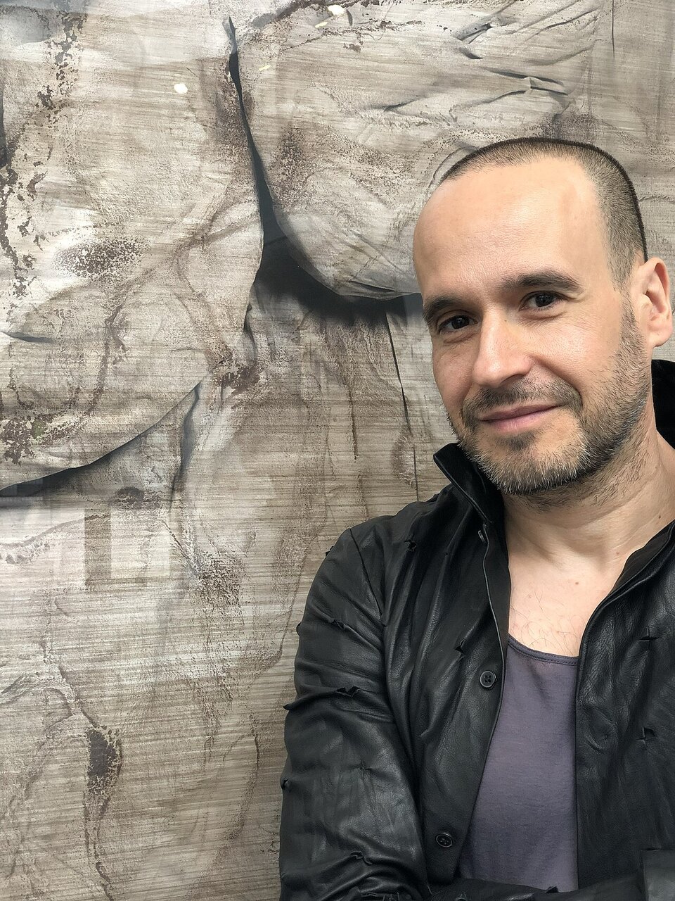 Kliknij żeby powiększyć Kolorowa fotografia przedstawiająca półpostaciowy portret młodego mężczyzny. Został on ukazany frontem do widza. Mężczyzna uśmiecha się i patrzy na wprost. Ma on wysokie, jasne czoło i krótko obcięte włosy z szerokimi zakolami. Jest ubrany w czarną, skórzaną kurtkę i granatowy podkoszulek. Tło stanowi betonowa ściana.