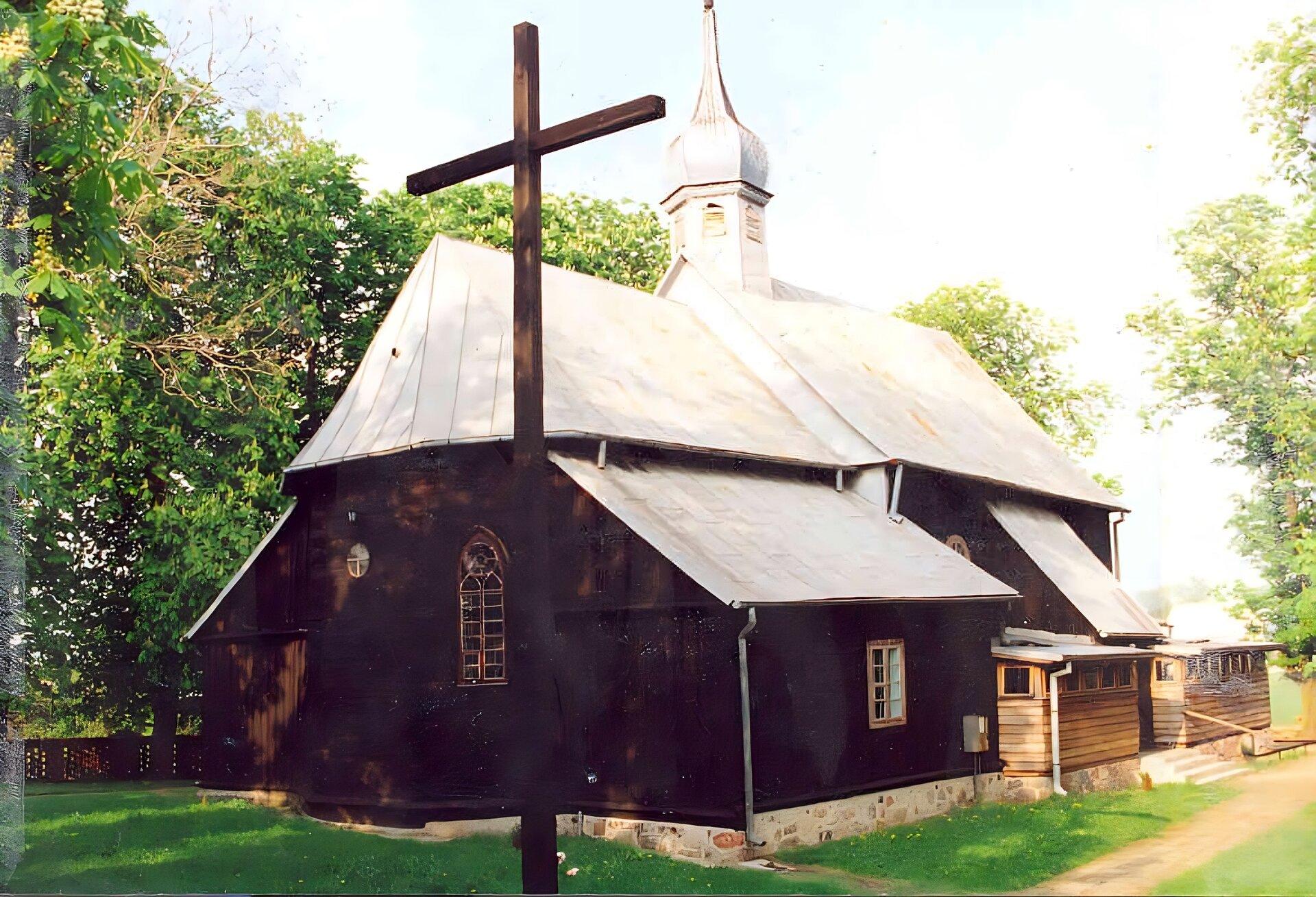Zdjęcie przedstawia czarny kościół od strony ołtarza. Ma on skośny, szary dach, na czubku którego znajduje się wieżyczka z kopułą. Przy kościele stoi krzyż w kolorze zbliżonym do kościoła. Wokół budynku rośnie zielona trawa, a po prawej stronie zdjęcia widoczne są zielone, wysokie drzewa. Zdjęcie zostało wykonane w świetle dziennym.