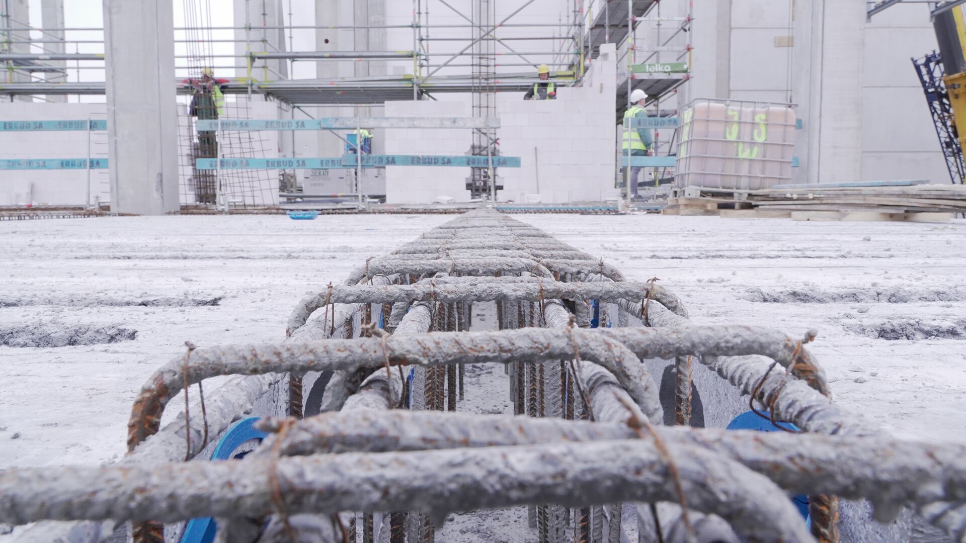 Zdjęcie przedstawia zbrojenie stropu pokryte białym pyłem. Za widoczną stalową konstrukcją można dostrzec dwóch robotników pracujących przy układaniu ściany.