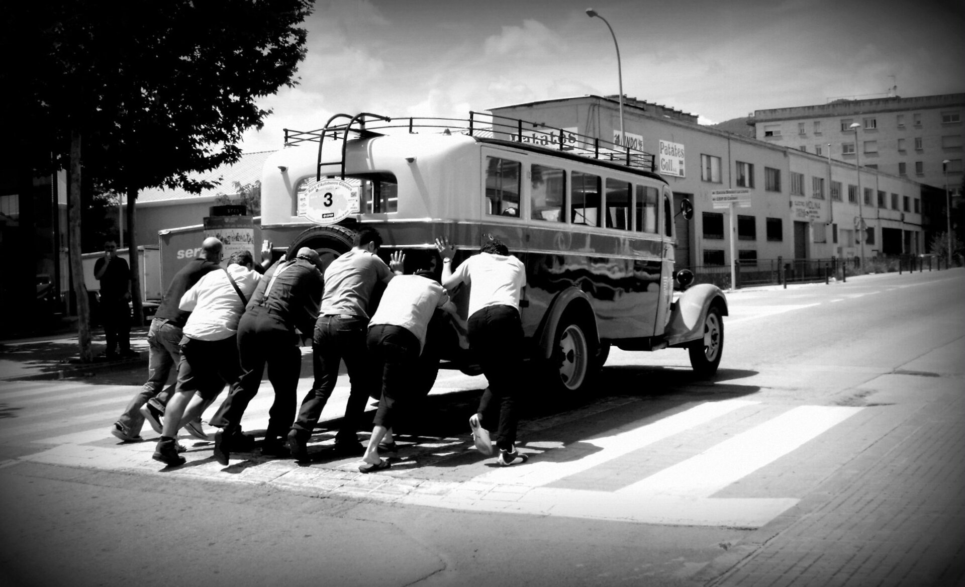 Czarno białe zdjęcie przedstawiające sześciu mężczyzn przepychających stary autobus ulicami małego miasteczka. Na budynkach w głębi kadru widoczne są hiszpańskojęzyczne napisy.