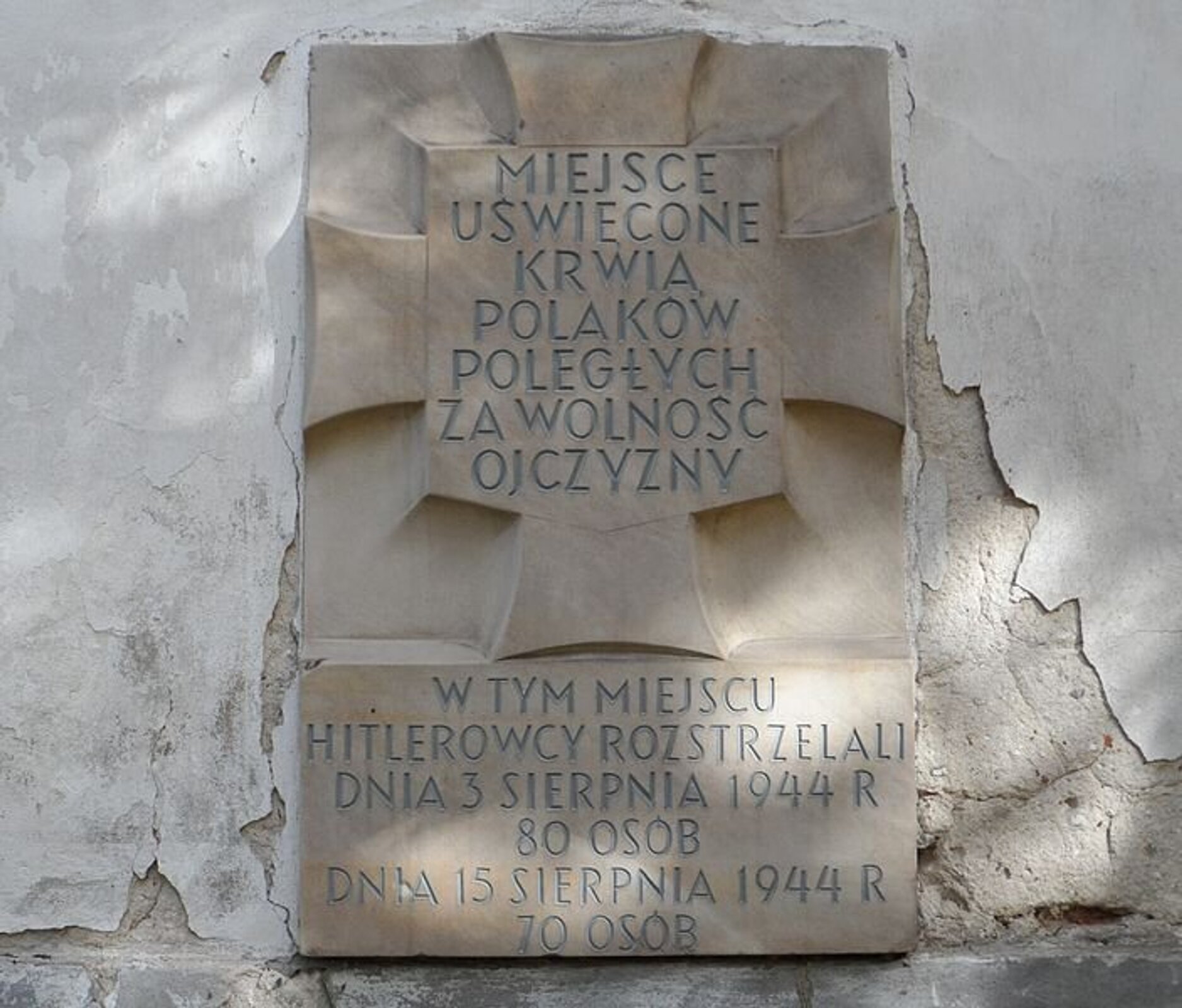Zdjęcie przedstawia na tle odrapanego z tynku muru kamienną tablicę, na której w centrum jest krzyż z planszą pośrodku i napisem: Miejsce uświęcone krwią Polaków poległych za wolność Ojczyzny. Poniżej krzyża z planszą na tablicy widnieje napis informacyjny: w tym miejscu hitlerowcy rozstrzelali dnia 3 sierpnia 1944 roku 80 osób, dnia 15 sierpnia 1944 roku 70 osób. 