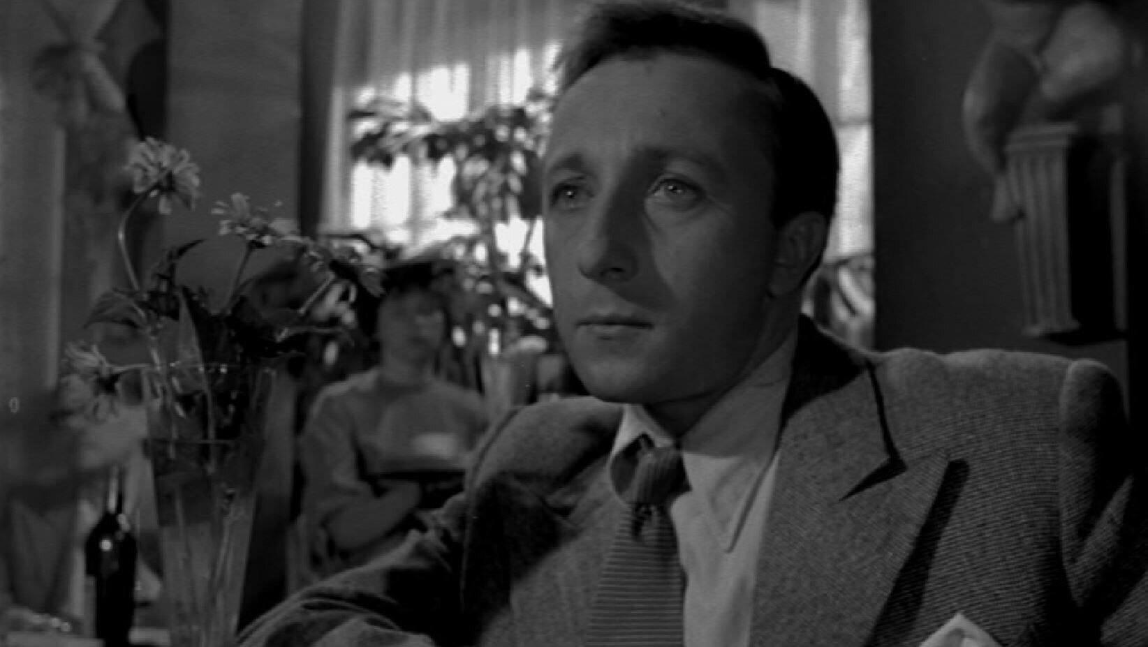 Zdjęcie przedstawia mężczyznę siedzącego przy stole, w kawiarni. Mężczyzna ubrany jest w garnitur. Jest zamyślony.