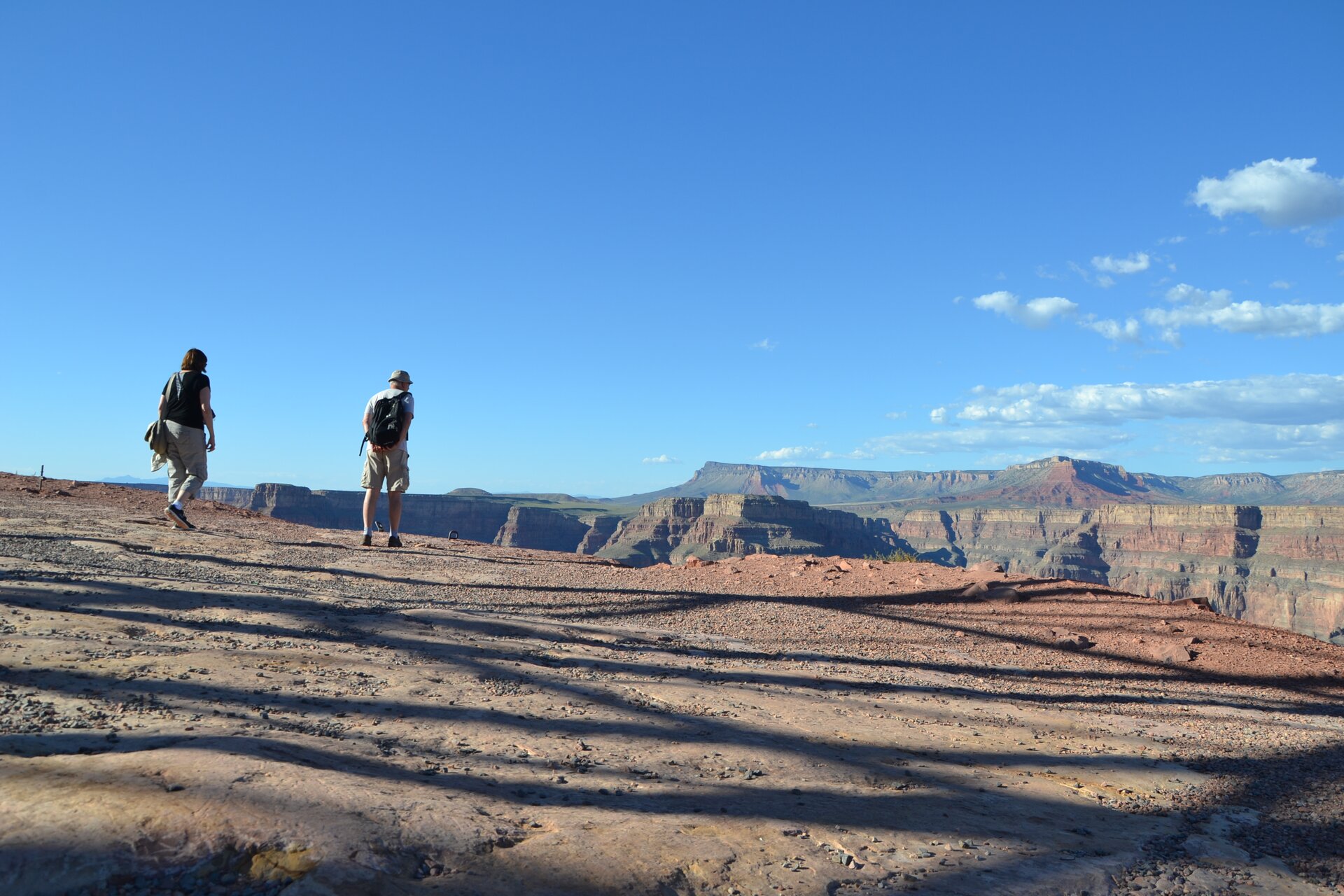 Fotografia przedstawia Wielki Kanion Kolorado w USA. Na ilustracji, z lewej strony znajdują się dwie idące postaci w kierunku kanionu. W oddali widać niemal równo zakończone pasmo górskie oraz błękitne niebo z małymi obłoczkami. Postaci idą po wielkiej płaskiej skale, na której jest wiele podłużnych cieni skrzyżowanych ze sobą.