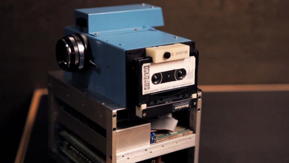 Ilustracja przedstawia fotografię aparatu cyfrowego. Aparat jest dużych rozmiarów, stoi na stole i sprawia wrażenie ciężkiego i masywnego. Ma kształt zbliżony do prostopadłościanu. Z jednej strony jest widoczny obiektyw, a drugiej kaseta magnetofonowa. 